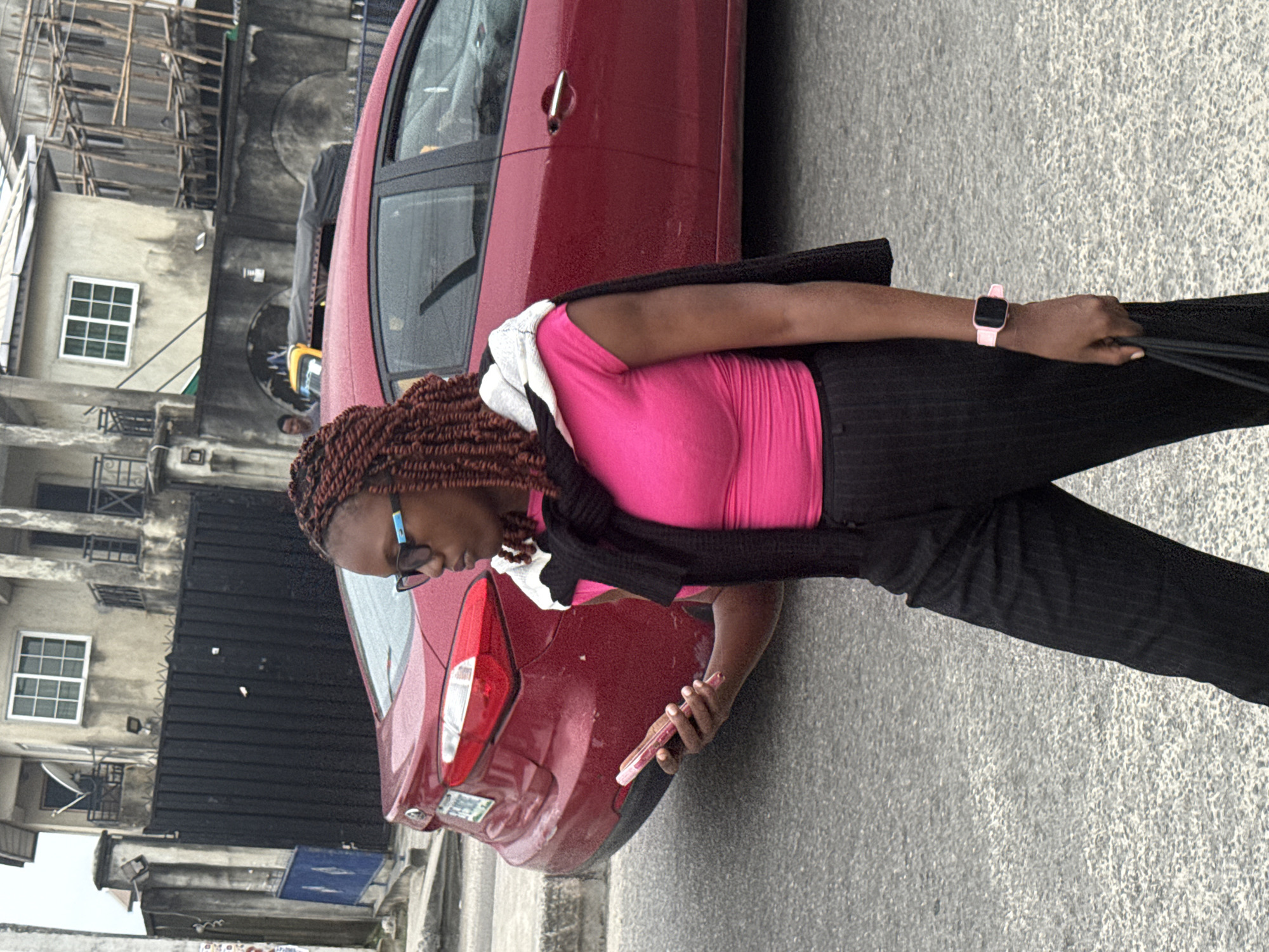 A woman with braided hair and glasses is walking on a street in Ado, Nigeria, looking at her phone. She is wearing a pink top, black striped pants, and a black sweater draped over her shoulders. A red car is parked on the street behind her, and in the background, there are buildings and a yellow vehicle. The pavement is asphalt, and the weather appears to be overcast.