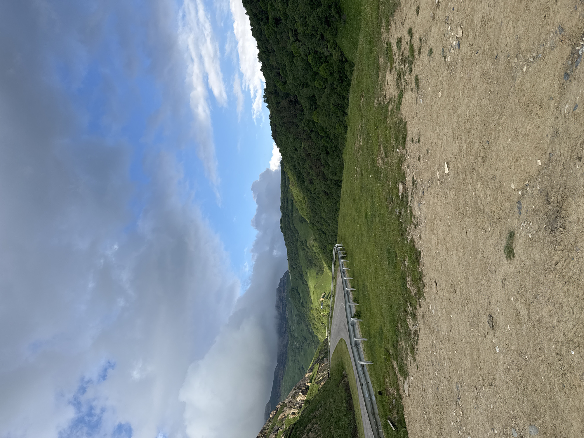 The image depicts a winding road through a lush green mountainous landscape under a dramatic sky. The road curves to the left, bordered by a metal guardrail. On the left side of the road, a steep, rocky mountain slope rises, partially obscured by clouds. To the right of the road, rolling green hills covered in dense forest stretch into the distance. The foreground is a dry, dusty patch of ground with scattered stones and sparse vegetation. The sky above is a mix of blue sky and heavy, dramatic clouds, with sunlight breaking through in some areas, illuminating the scene. The location is identified as Verkhnyaya Mara, Russia. There are no people or animals visible in the image, and no explicit actions are taking place. The scene is a natural landscape with a man-made road.