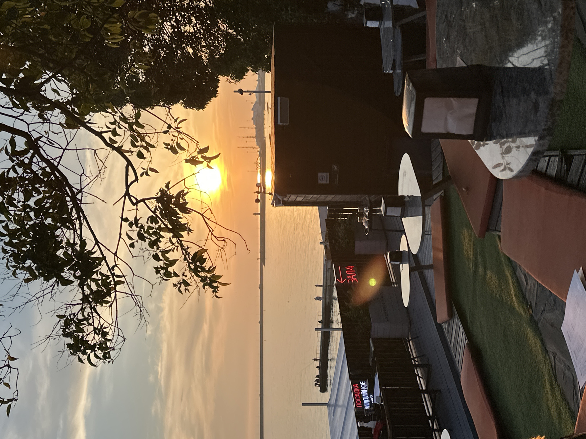 This photograph captures a sunset scene overlooking a body of water, likely the Black Sea, from an elevated outdoor cafe or restaurant terrace in Sochi, Russia. The sun, a bright orange orb, is positioned low on the horizon, casting a warm, golden glow across the sky and reflecting on the calm water surface. Silhouetted tree branches and leaves frame the top and right portions of the image.
In the mid-ground, a long pier or breakwater extends into the water, with several individuals visible, most of whom appear to be fishing. Further in the distance, on the horizon, multiple tall structures resembling ship masts, cranes, or industrial elements of a port are visible, along with what could be a lighthouse or a beacon on a distant landmass.
The foreground depicts the outdoor terrace of a dining establishment. On the left, a white canopy covers a section of the seating area. Below it, a counter holds a spray bottle. Neon signs in Russian are visible: one red sign reads "ПОСАДКА МЛАДИМНОЕ" (Seating Mladimnoe) and another, also red, points left with an arrow and the word "ЗАЛА" (Hall/Room). The terrace features dark wooden decking mixed with sections of artificial green turf. Various seating arrangements are present, including tables with dark, possibly marble, tops and chairs or low benches with orange cushions. A dark, rectangular utility structure or small building stands on the right side of the terrace, with a weather vane (anemometer) mounted on its roof. Napkin dispensers are visible on some of the tables. The overall ambiance is tranquil, illuminated by the fading daylight.