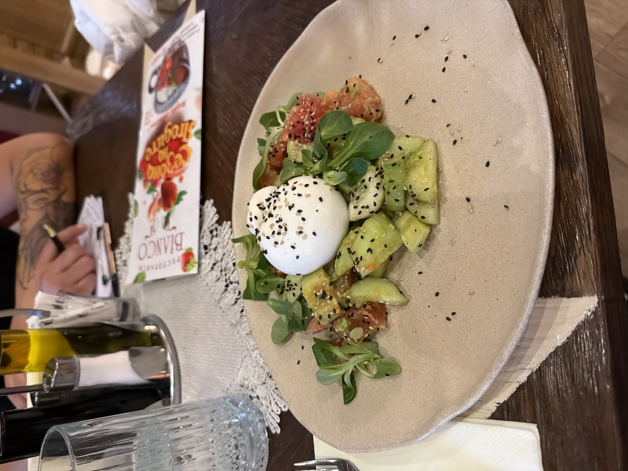 The image depicts a plate of food on a wooden table, likely in a restaurant. The dish consists of a white ball of mozzarella-like cheese, surrounded by chopped cucumbers, tomatoes, and green leaves (likely lettuce or lamb's lettuce). Black sesame seeds are sprinkled over the cheese and scattered around the plate. Part of a restaurant menu is visible, showing the name "Bianco" and something written in Bulgarian. Additionally, part of a tattooed arm holding a pen and a rack with a glass and bottles of oil is partially visible. The location is assumed to be Veliko Tarnovo, Bulgaria based on the provided information.