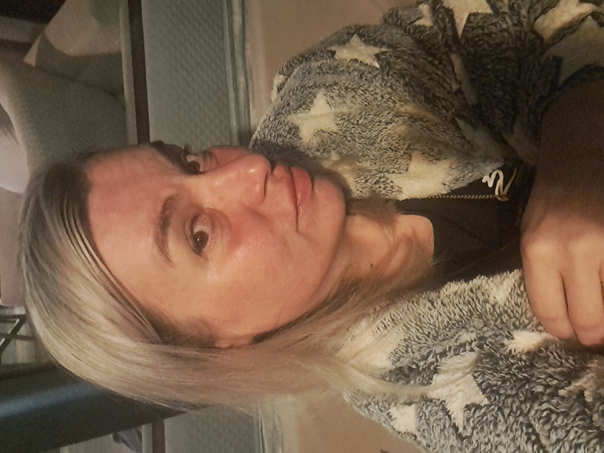 The image shows a medium close-up of a woman with blonde hair and fair skin, wearing a fuzzy blanket with a star pattern. Her head is tilted and she is looking at the camera with a slightly open mouth. The background is out of focus and appears to be an indoor setting with furniture. There is no information available about the specific location within Krasnyi Ogorok, Russia.