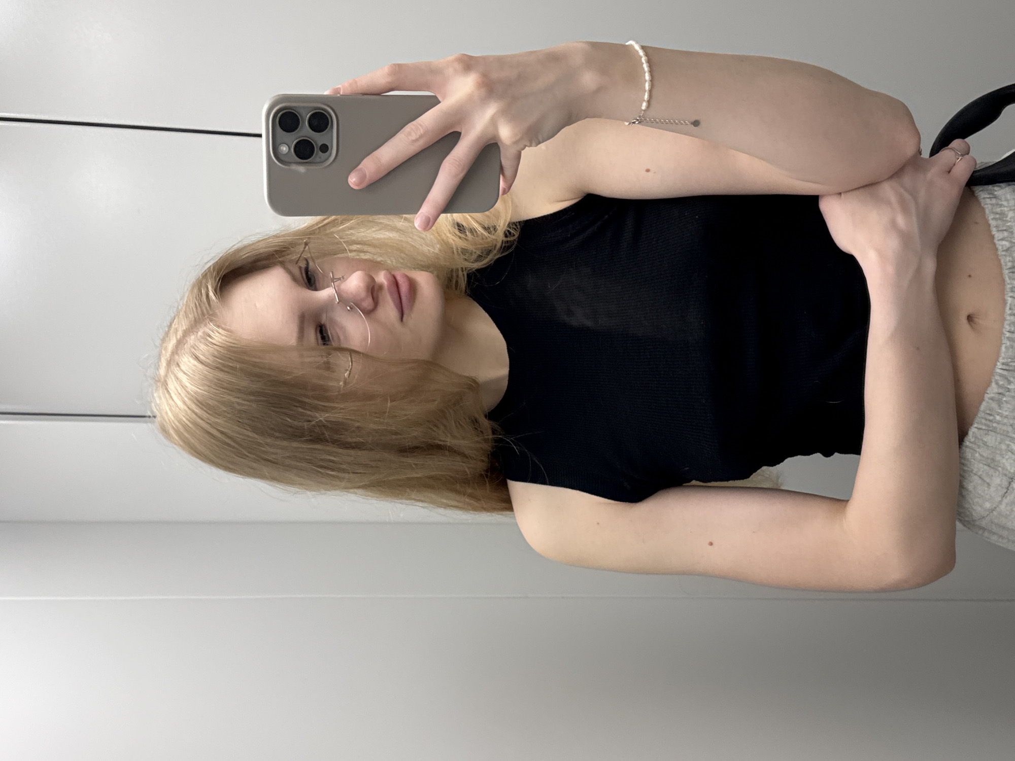 In a mirrored elevator, a young woman with long blonde hair and wire-rimmed glasses takes a selfie. She is wearing a black tank top and grey sweatpants. On her left wrist, she wears a pearl bracelet. The elevator walls are plain grey. The image does not contain information that allows for identification of the city of Perm, Russia.