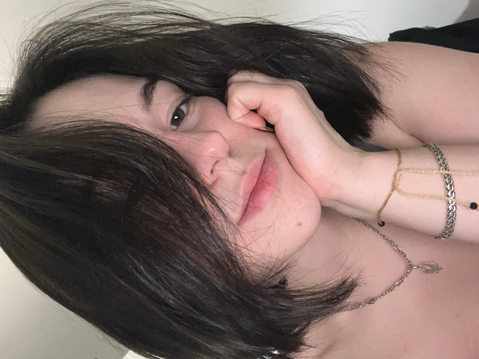 A woman with short, dark brown hair is resting her head on her hand, close to the camera. She is wearing a silver necklace with a pendant and two bracelets on her wrist: one gold and one silver. Her skin appears fair, and her gaze is soft. The photo has a casual, intimate feel.