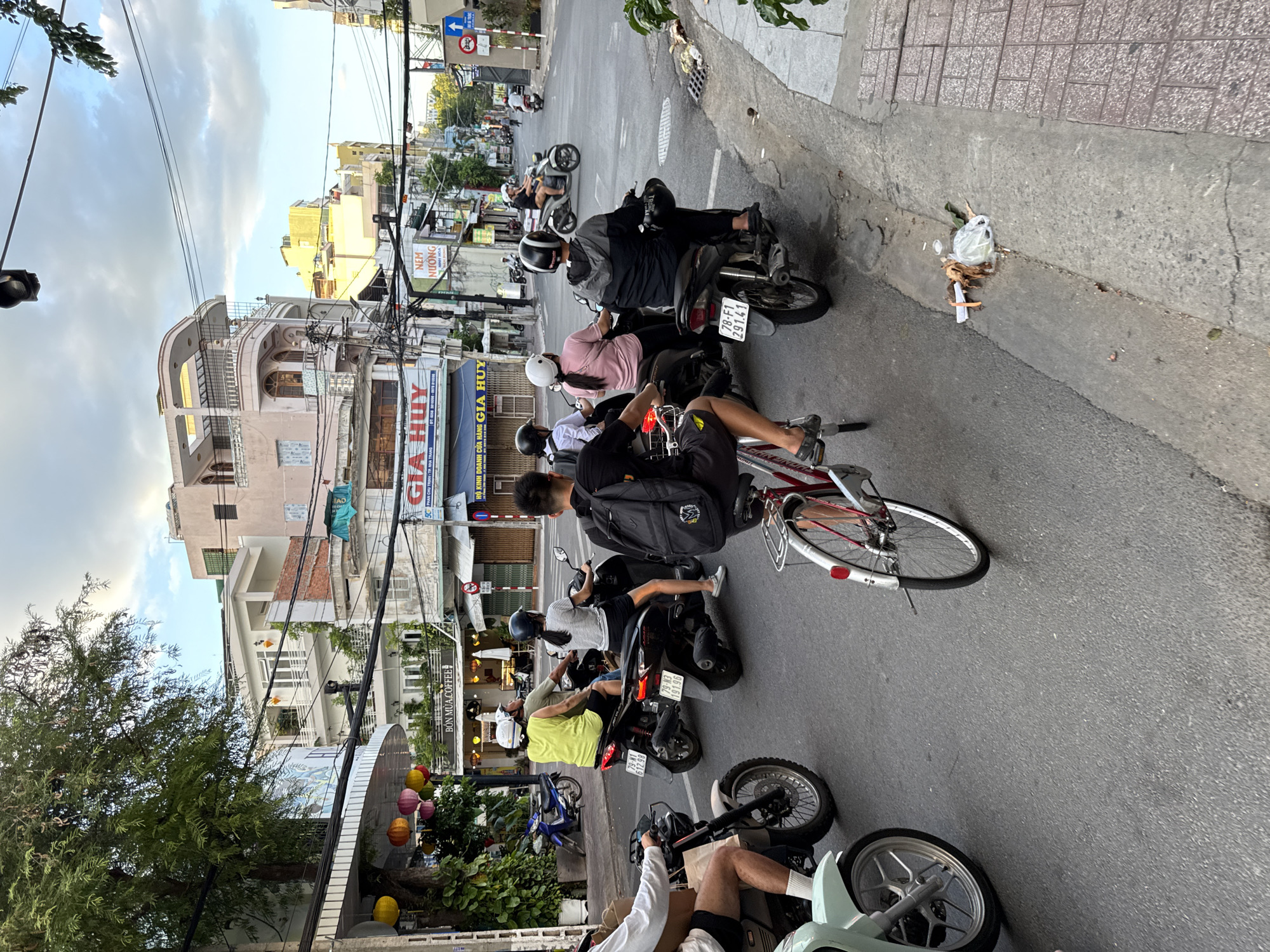 In a street scene in Nha Trang, Vietnam, several people are riding motorcycles and bicycles. The day is partly cloudy, with buildings lining both sides of the street. On the left, a large tree with lush foliage shades a sidewalk lined with paper lanterns in yellow, pink, and orange. Several shops are visible, including "Bôn Mùa Coffee". A blue motorcycle is parked on the side. In the center of the image, a person in a bright yellow shirt and white helmet rides a motorcycle, followed by a person in a striped shirt and black helmet. Another motorcycle is ridden by a person in a grey hooded jacket. In the foreground, a person rides a bicycle with a red frame and a person on a light blue scooter is also visible. The street has clear lane markings. On the right side of the street, a sidewalk is visible with some trash on the curb. Traffic signs, including a directional arrow, are also present. The buildings have a mixture of architectural styles, with balconies and potted plants. The overall impression is a busy urban street scene in Vietnam.