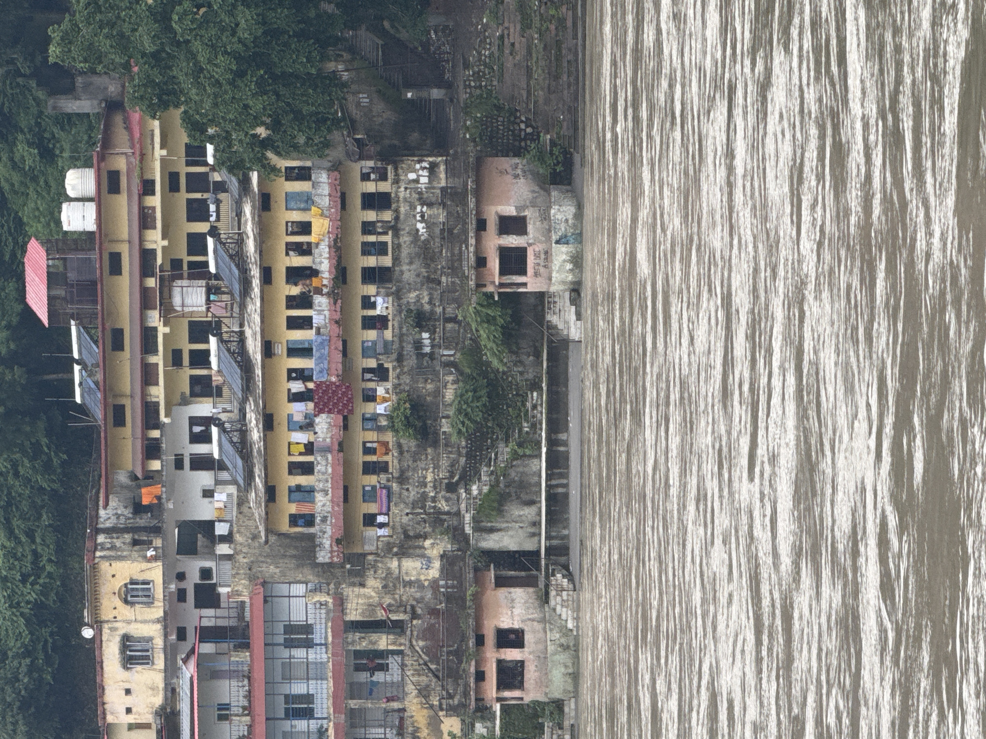 The image captures a multi-story building situated on the banks of the Ganges River in Rishikesh, India. The architecture features a pale yellow facade with multiple balconies adorned with laundry hanging out to dry. Solar panels are visible on the rooftops of some sections. In the foreground, the Ganges River flows with visible ripples, reflecting the overcast sky. At the base of the building, a small, weathered pink structure with two windows and graffiti on its exterior sits close to the riverbank, accessible by stone steps. The overall scene depicts a densely populated urban environment adjacent to a significant natural water body.