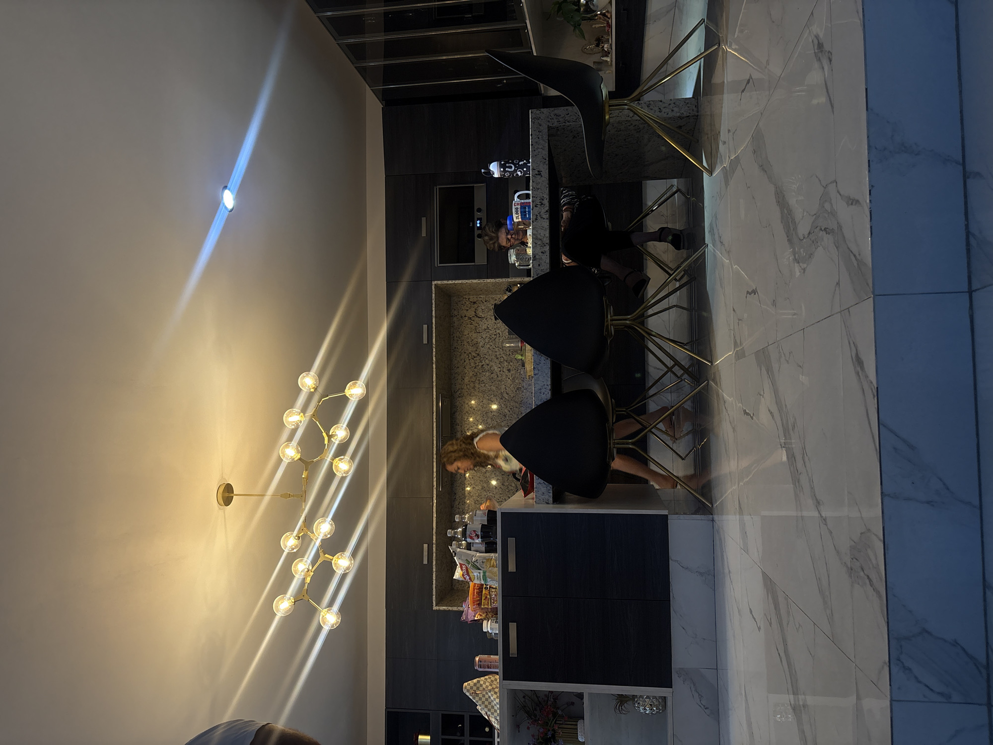 A brightly lit, modern interior in San Bernardino Tlaxcalancingo, Mexico, features a kitchen with dark cabinetry, a light-colored countertop, and bar stools with gold-colored legs. Two women are present, one seated at the counter and another standing near the lower cabinets. Various food items and beverages are visible on the counter and within the cabinetry. A chandelier hangs from the ceiling, casting light patterns on the wall. The flooring is light-colored with marble-like veining. A section of blue tile is on the right side of the image.