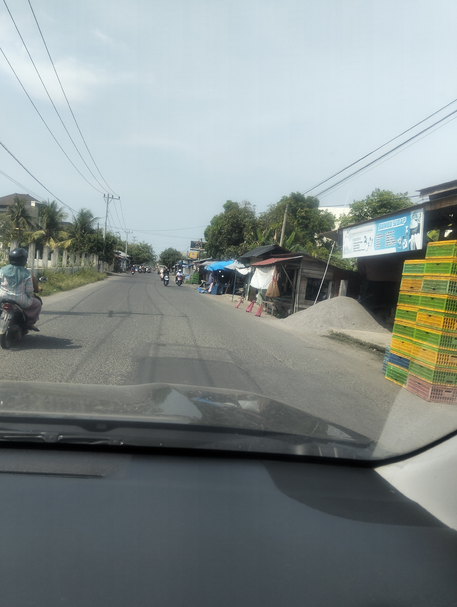 The image depicts a street scene in Geulanggang Teungoh, Indonesia, taken from inside a vehicle, looking out through the windshield. The road surface is asphalt with visible tire marks. On the left side of the road, a person wearing a helmet and a light-colored patterned shirt and dark pants is riding a scooter away from the camera. Further down the road on the left, a fence and palm trees are visible. On the right side of the road, there are several small market stalls or shops. One stall has a pile of sand next to it, and another has stacks of colorful plastic crates, predominantly green, yellow, and pink, and a sign that reads "BUMBU SEDAP". Several other motorcycles are seen in the distance on the road. Utility poles and power lines are present overhead. The sky is overcast with a light blue hue.