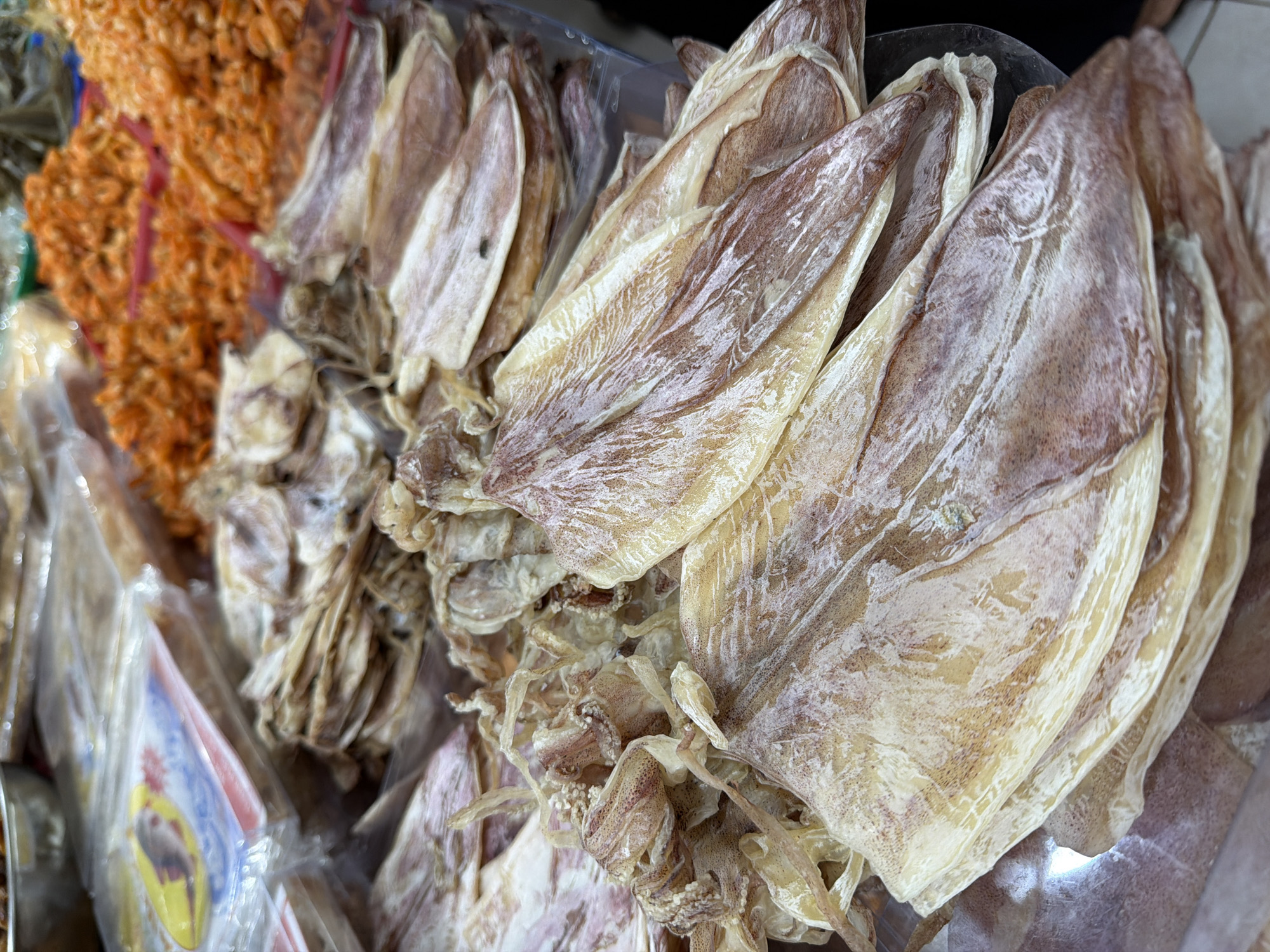 The image displays a close-up view of dried seafood, specifically dried squid and dried shrimp, at a market stall in Nha Trang Ward, Vietnam. The foreground is dominated by a pile of large, flattened, dried squid. These squid vary in color from pale yellow to light brown with some reddish-brown markings. Their texture appears leathery and translucent in places. To the right and in the background, smaller, bright orange dried shrimp are piled in red plastic containers. Some plastic packaging with colorful labels is visible behind the squid. The overall impression is one of a food market offering dried seafood products.