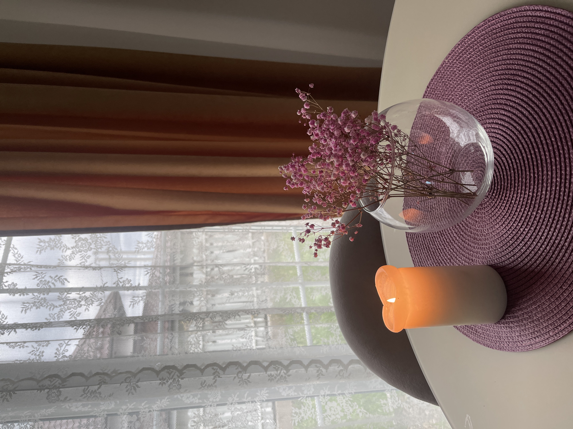 A high-angle shot depicts a scene indoors, likely near a window. The composition features a white table with a round, purple woven placemat. Resting on the placemat is a lit, cylindrical candle with an orange glow. Beside the candle is a clear glass vase containing pink baby's breath flowers. In the background, a window covered with white lace curtains is visible. Part of a chair's back is also noticeable, and brown drapes are visible at the top of the frame. The location of Хлябово, Russia is not evident from the image.