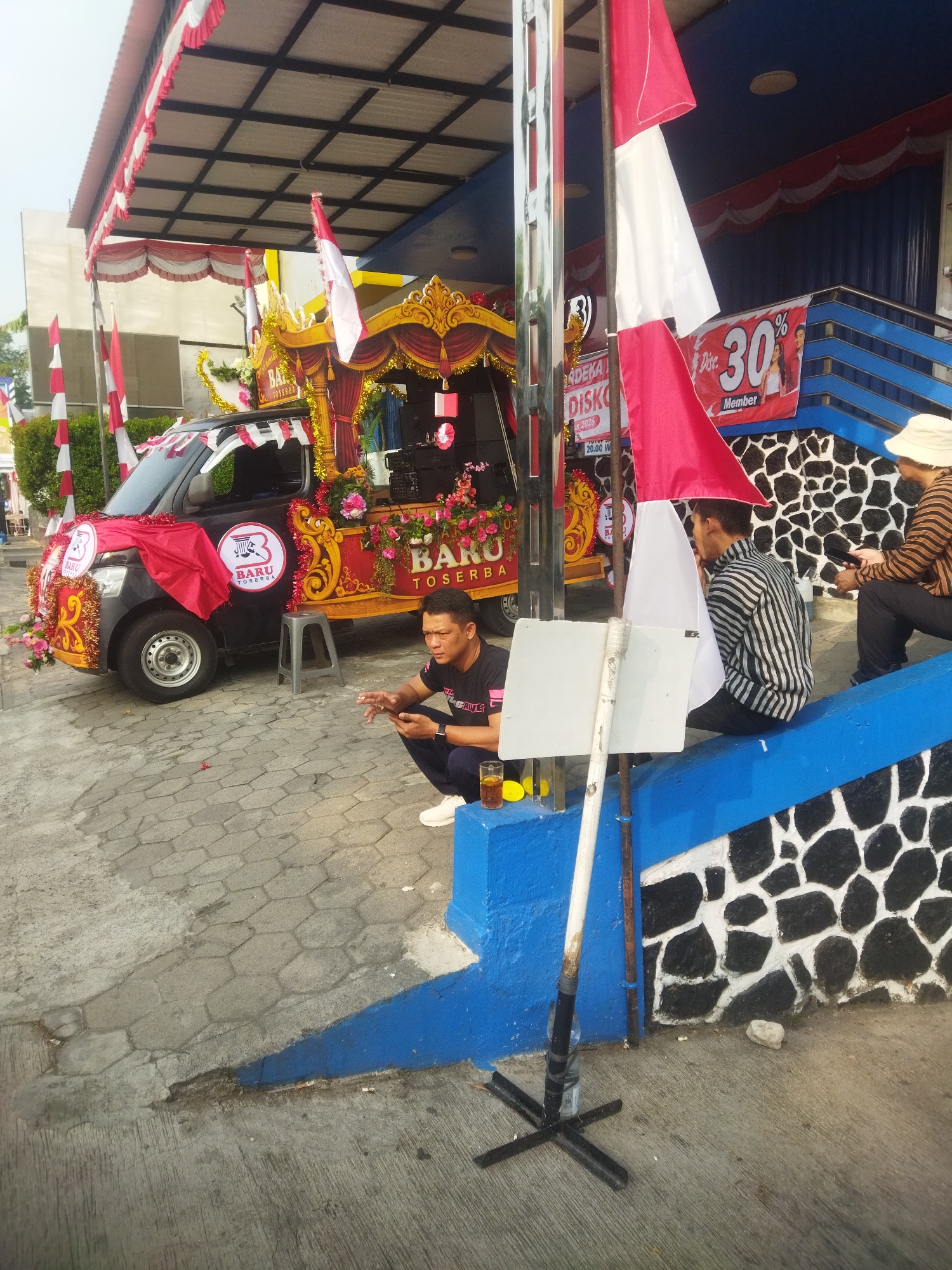 The image depicts a street scene in the city of Wonogiri, Indonesia, featuring a decorated pickup truck that appears to be part of a promotional or celebratory event. The truck is adorned with red and white flags, floral arrangements, and gold trim, with the logo "BARU TOSERBA" prominently displayed. Several people are present. In the foreground, a man in a black t-shirt and blue pants is crouching and looking at his phone, with a glass of iced tea beside him. To his right, another man in a striped shirt is sitting on a blue-tiered structure, also looking at his phone. Further to the right, a person wearing a hat and a striped shirt is sitting on the same structure, holding a phone. The background shows a building with blue and white striped siding, along with more red and white flags and banners, one of which advertises a "30% Discount". The ground is paved with hexagonal stones. The overall atmosphere suggests a public gathering or event taking place outdoors during the daytime.