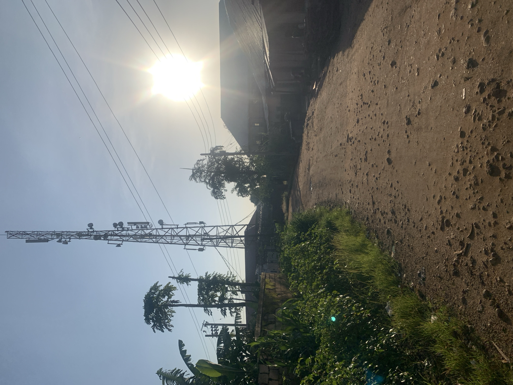 In Rafin Par, Nigeria, a dirt road leads towards a large cell tower under a bright, hazy sun. The road is unpaved and rough, with scattered stones and debris. To the left of the road, lush green vegetation and banana trees grow along a low stone wall. In the background, to the right of the cell tower, are several buildings with corrugated metal roofs, suggesting a residential or commercial area. The sky is clear but washed out by the sun's glare, indicating it is likely morning or late afternoon. No people are visible in the image.