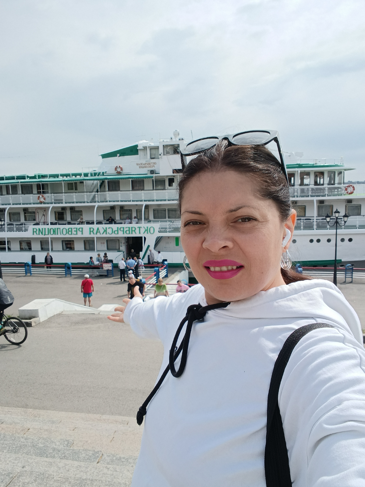 A woman takes a selfie in the foreground, pointing towards a large white and green river cruise ship docked behind her. She has dark hair pulled back, with sunglasses resting on her head. She wears a white hooded sweatshirt with black straps from a bag, bright pink lipstick, hoop earrings, and a white earbud in her right ear. Her expression is pleasant as she looks towards the camera.
The multi-deck ship, likely a passenger vessel, features numerous windows, railings, and green trim. Prominently displayed on its side are large green letters that appear reversed in the selfie, spelling out "RNIUIONOPEВЯ RAXJPARTKO". When read correctly, this text translates from Russian as "ОКТЯБРЬСКАЯ РЕВОЛЮЦИЯ" (Oktyabr'skaya Revolyutsiya), meaning "October Revolution." People are visible on the ship's lower deck and on the concrete pier, suggesting boarding or disembarking. A gangway with blue and red barriers connects the ship to the dock.
In the mid-ground, a person in a red shirt stands near the barriers, and to the far left, part of a bicycle is visible. The setting is an outdoor paved dock or riverbank, under a bright, overcast sky. The scene depicts a lively daytime atmosphere at a port, likely in Saratov, Russia, as implied by the ship's name and given context.