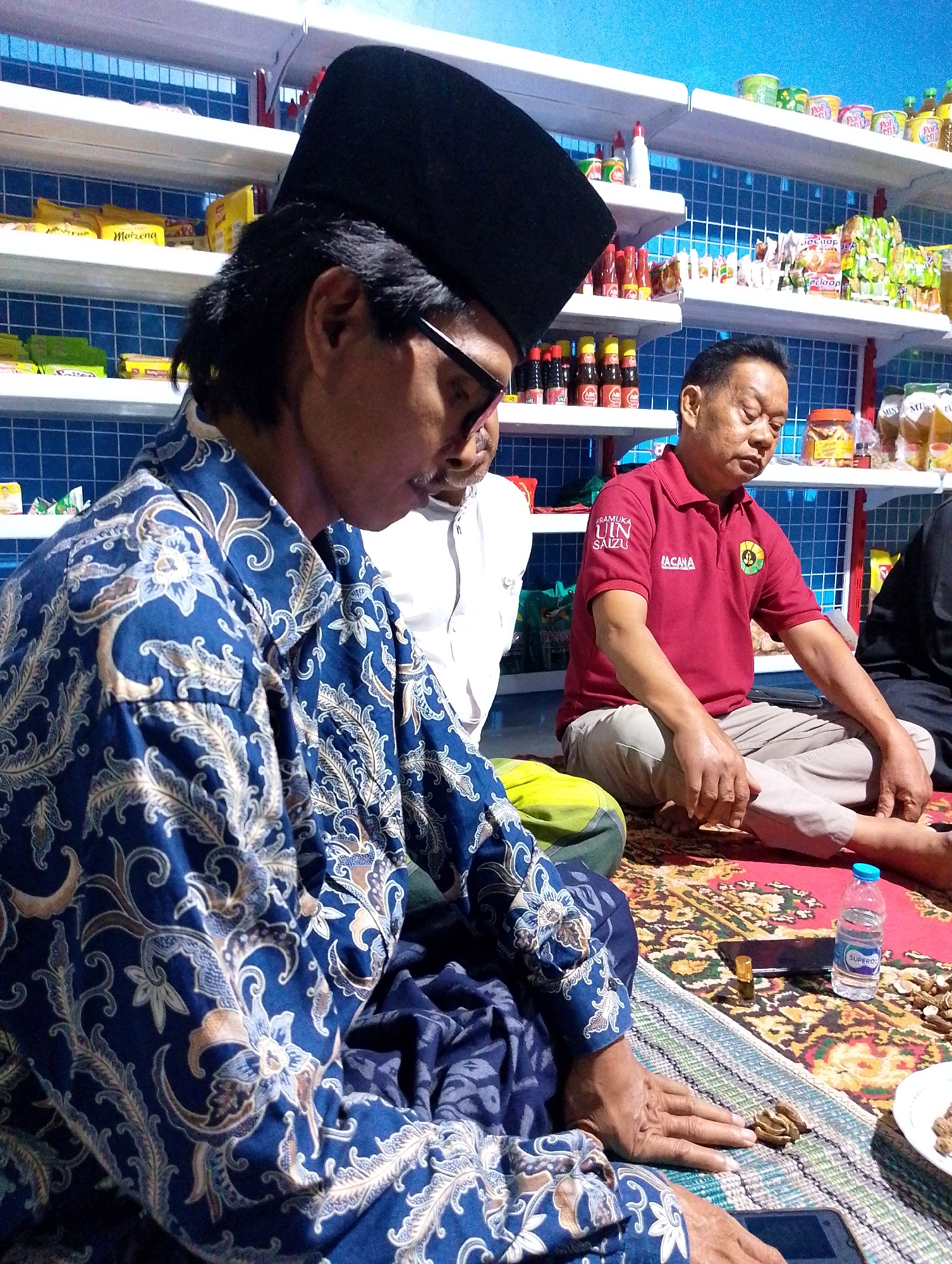 Three men are sitting on the floor in what appears to be a small convenience store or warung in Cilacap, Indonesia. The man in the foreground on the left is wearing a blue batik shirt and a black peci (skullcap). He has glasses and is looking down. To his right, a man in a red polo shirt with writing on it and khaki pants is also seated. He has his eyes closed and appears to be resting. A third man, mostly out of frame on the far right, is wearing a dark shirt. The men are sitting on a rug with a woven mat placed on top of it in front of them. On the mat, there are small food items, a bottle of Super O2 water, and a small golden vial. In the background, shelves are stocked with various packaged goods, including bottles of sauces, snacks, and other retail items. The walls are painted blue. The scene suggests a casual gathering or meeting within a commercial space.
