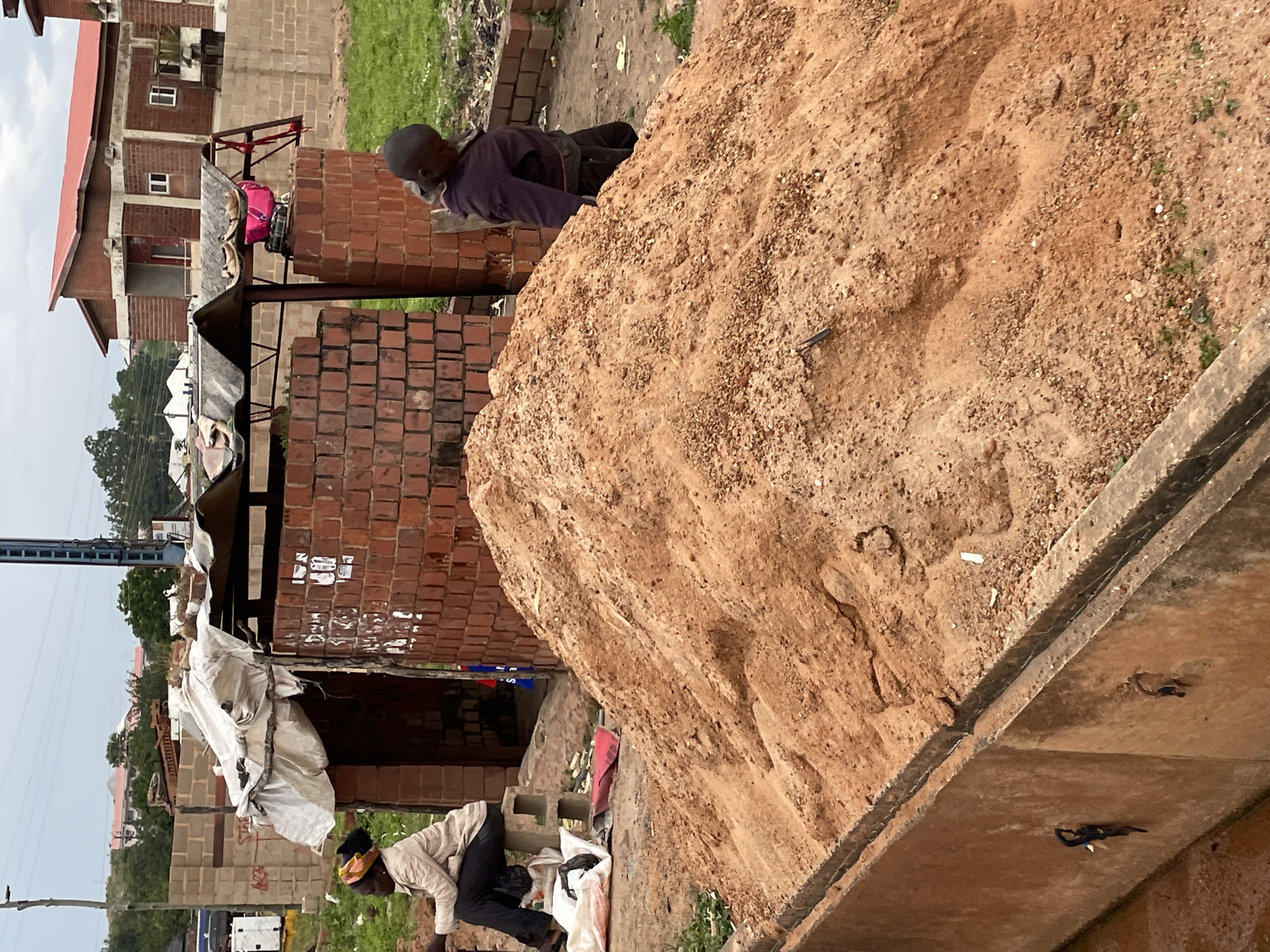 In Jos, Nigeria, a scene unfolds featuring a large pile of orange sand in the foreground, possibly for construction. To the left, a woman sits on a concrete block, wearing a light-colored jacket and dark pants, her head adorned with a yellow and black headwrap. She appears to be resting or observing the surroundings. To the right of the sand pile, a young person in a dark purple jacket and dark pants stands, looking downwards. Behind the sand pile, a simple structure with a corrugated metal roof, supported by brick walls, is visible. The overall environment suggests an outdoor setting, likely a construction site or an area undergoing development, with buildings and vegetation in the background under an overcast sky.