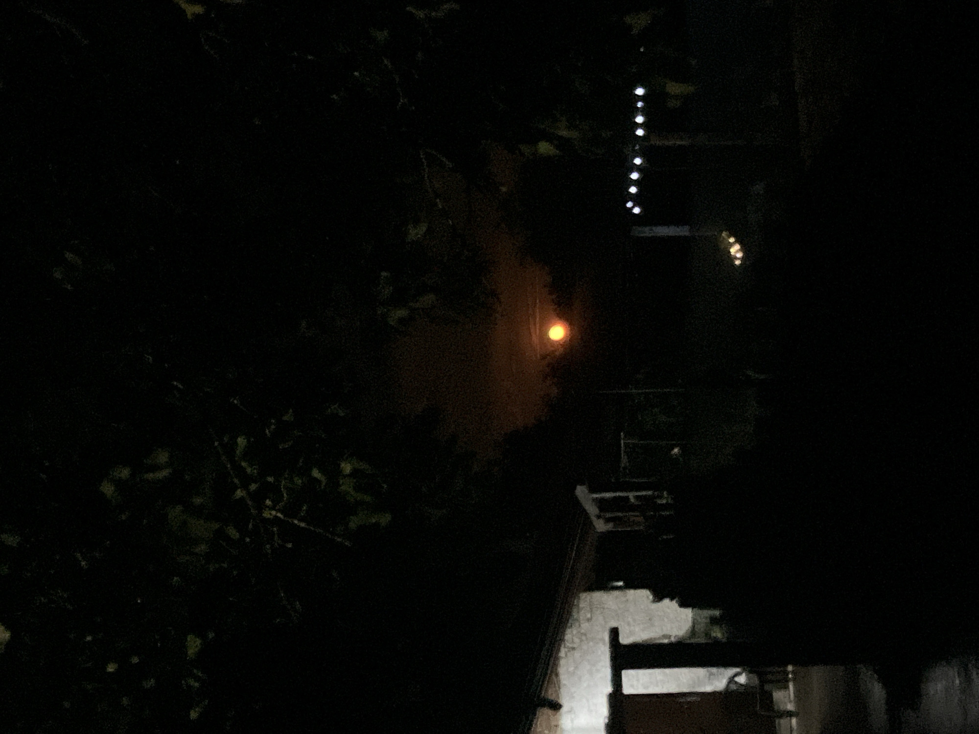 The image depicts a dimly lit outdoor scene at night, with the sky dominated by darkness and a faint orange glow in the upper center, possibly indicating a moon partially obscured by clouds or atmospheric conditions. In the foreground, a portion of a building, likely a house, is visible on the left, with a light source illuminating its exterior wall. The structure appears to be made of rough-hewn materials. To the right of the building, the landscape is obscured by shadows and foliage from overhanging trees. In the background, a line of small, bright lights, potentially decorative string lights, is visible against the dark backdrop, suggesting a garden or outdoor living area. The overall impression is one of a quiet, rural or suburban setting at night. The location is identified as Kramolin, Bulgaria. There are no visible people or animals in the image.