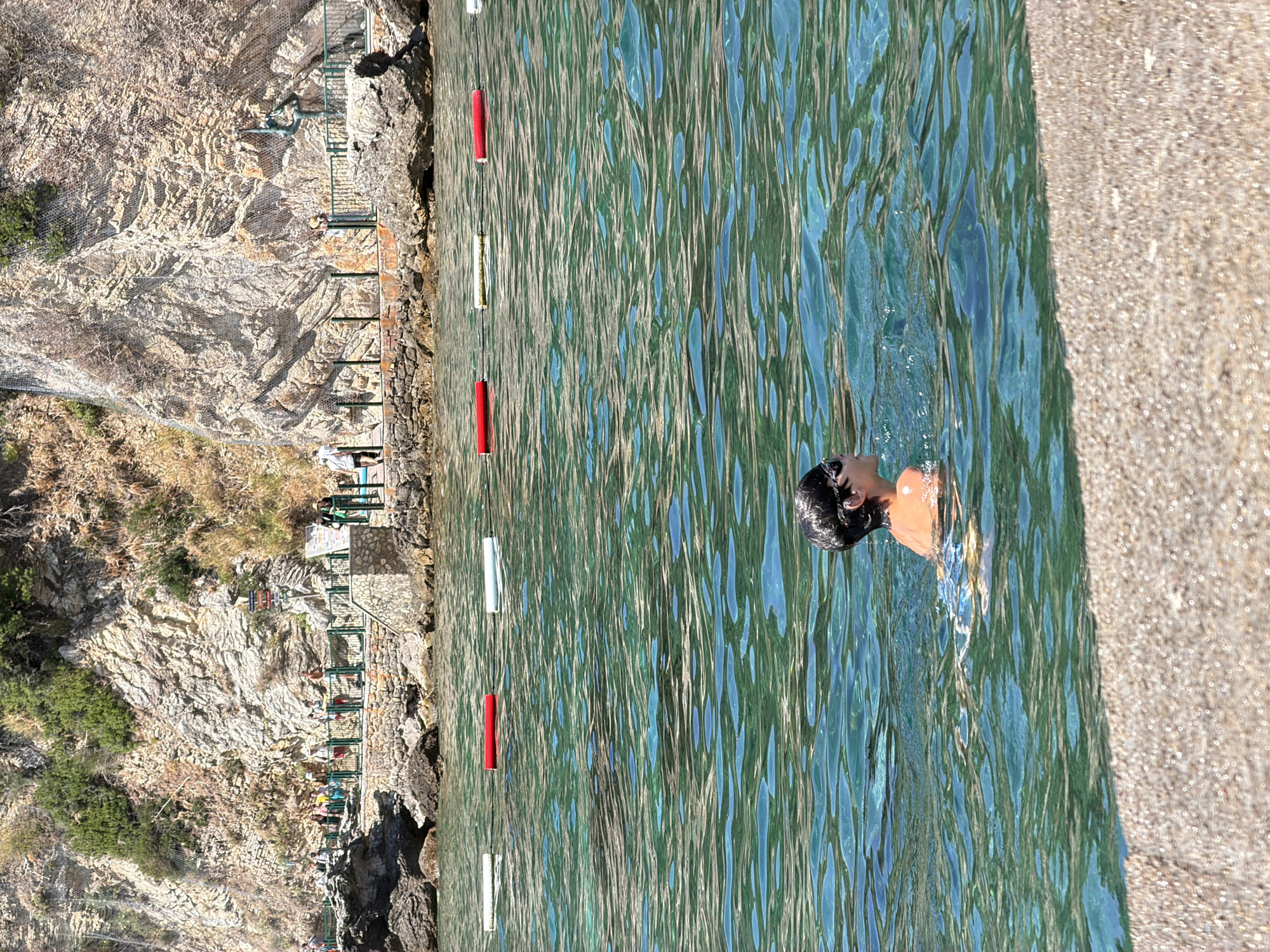 The image captures a scenic view of a coastal area in Budva, Montenegro. In the foreground, a young boy is swimming in the clear, turquoise waters of the Adriatic Sea. He is wearing swimming goggles and appears to be enjoying his time in the water. The water is slightly rippled, reflecting the bright sunlight. A string of red and white buoys marks a section of the water.
In the middle ground, a rocky shoreline is visible, with a stone pathway and a metal fence running along it. Several people are strolling along the path, enjoying the coastal view. Behind the path, a steep, rocky cliff face rises, covered in sparse vegetation. The cliff shows evidence of erosion and geological stratification.
The background depicts a wider view of the coastline, with more rocky outcrops and lush greenery on the hillsides. The overall impression is one of a beautiful, sunny day at a popular tourist destination. The architectural elements, such as the stone pathway and metal fence, suggest a developed tourist area, likely a beach or promenade. The presence of people indicates that this is an active and frequented location.