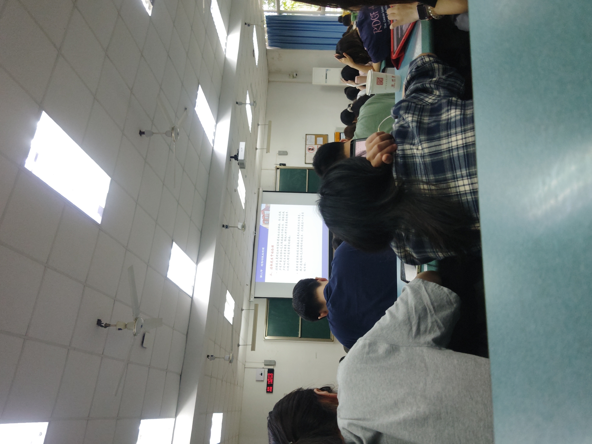 Here is a detailed description of the image:
**Scene Overview:** The image captures a classroom setting, likely a lecture or presentation, in Chongqing High-tech Zone, China. The room is well-lit with fluorescent ceiling lights. Multiple rows of students are seated, facing towards the front of the room where a projection screen displays text and graphics related to "Eugenics ethics". The perspective is from one of the desks, looking slightly down and across the room.
**People:** Students are the primary subjects. Their heads, shoulders, and backs are visible, suggesting they are engaged in listening or taking notes. There are men and women in the room, some wearing casual attire. One student is wearing a plaid shirt, another has on a gray sweatshirt. The general impression is of an attentive audience.
**Objects:**
* **Seating:** Student desks are visible.
* **Projection Screen:** The screen shows the presentation material, with Chinese characters and what appears to be subheadings.
* **Ceiling Lights:** Long fluorescent lights are arranged in rows.
* **Ceiling Fans:** A ceiling fan is noticeable on the ceiling.
* **Wall Clock**: A wall clock with LED display shows time.
* **Window**: A window is on the side of the wall.
**Actions:** Students are seated, presumably listening to a lecture.
**Environment:** The room appears clean and organized, with a standard classroom layout. The lighting is bright, and the overall atmosphere suggests a formal learning environment.