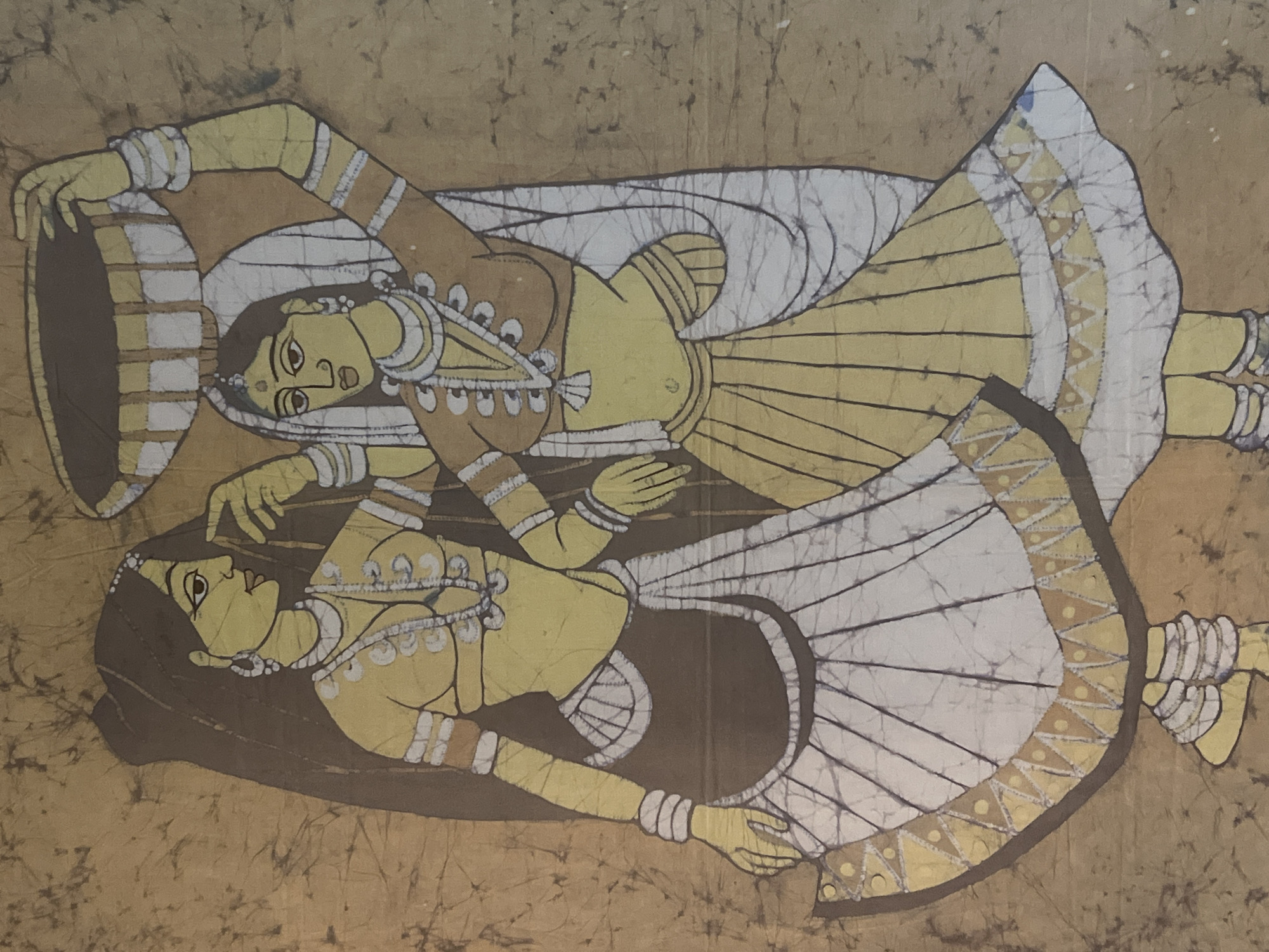 The image is a batik painting depicting two women in traditional Indian clothing. Both are adorned with jewelry and are wearing skirts and tops. The background has a crackled texture. The location is not discernible from the image.