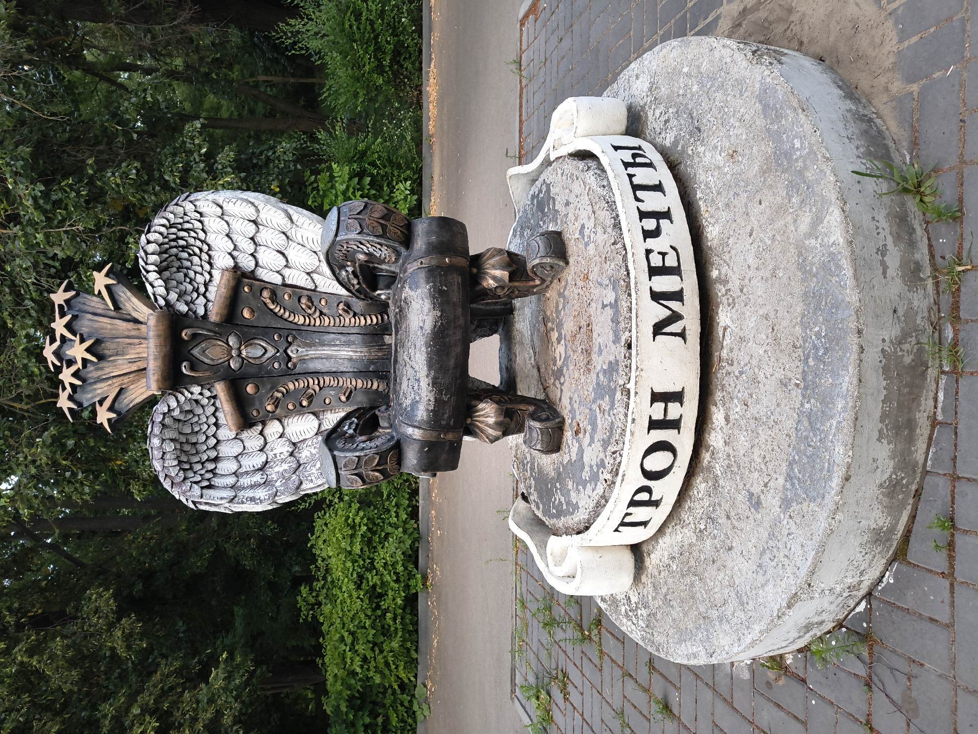The image depicts a large, ornate throne-like sculpture situated outdoors. The sculpture is dark-colored with metallic accents and features elaborate wings on either side, resembling feathers. At the top, it is adorned with golden star-like shapes. The sculpture is mounted on a weathered concrete pedestal. A white band encircles the pedestal, featuring black Cyrillic lettering that reads "ТРОН МЕЧТЫ," which translates to "Throne of Dreams." The sculpture is placed on a paved area made of gray bricks, surrounded by greenery and trees, suggesting a park or public space. The background shows dense foliage and a pathway. There are no people visible in the image. This sculpture is located in the city of Mikhaylovka, Russia.