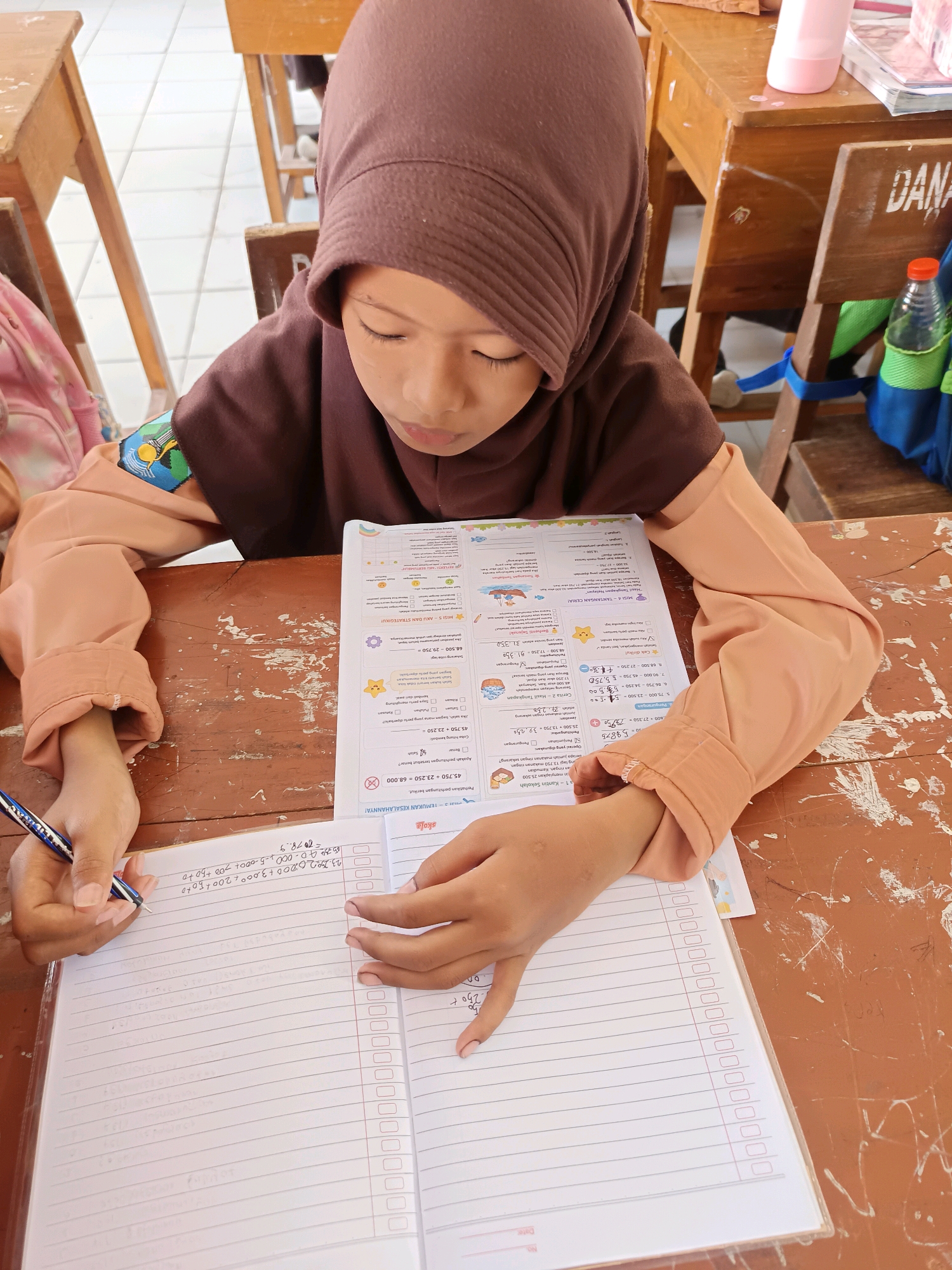 A young girl wearing a brown hijab and peach-colored long-sleeved shirt is seated at a wooden desk, writing in a notebook. She is focused on her work, with her eyes directed downwards. The desk surface is worn and stained, typical of school furniture in Indonesia. The background shows other wooden desks and chairs, suggesting a classroom setting. On the desk, a workbook with colorful illustrations is open, and a blue pen is in the girl's hand. Her left hand rests on the workbook, and a ring is visible on her right ring finger. The scene is well-lit, likely by natural light from a window. The image appears to be taken in a classroom in Labuhan, Indonesia, judging by the visual cues and context.