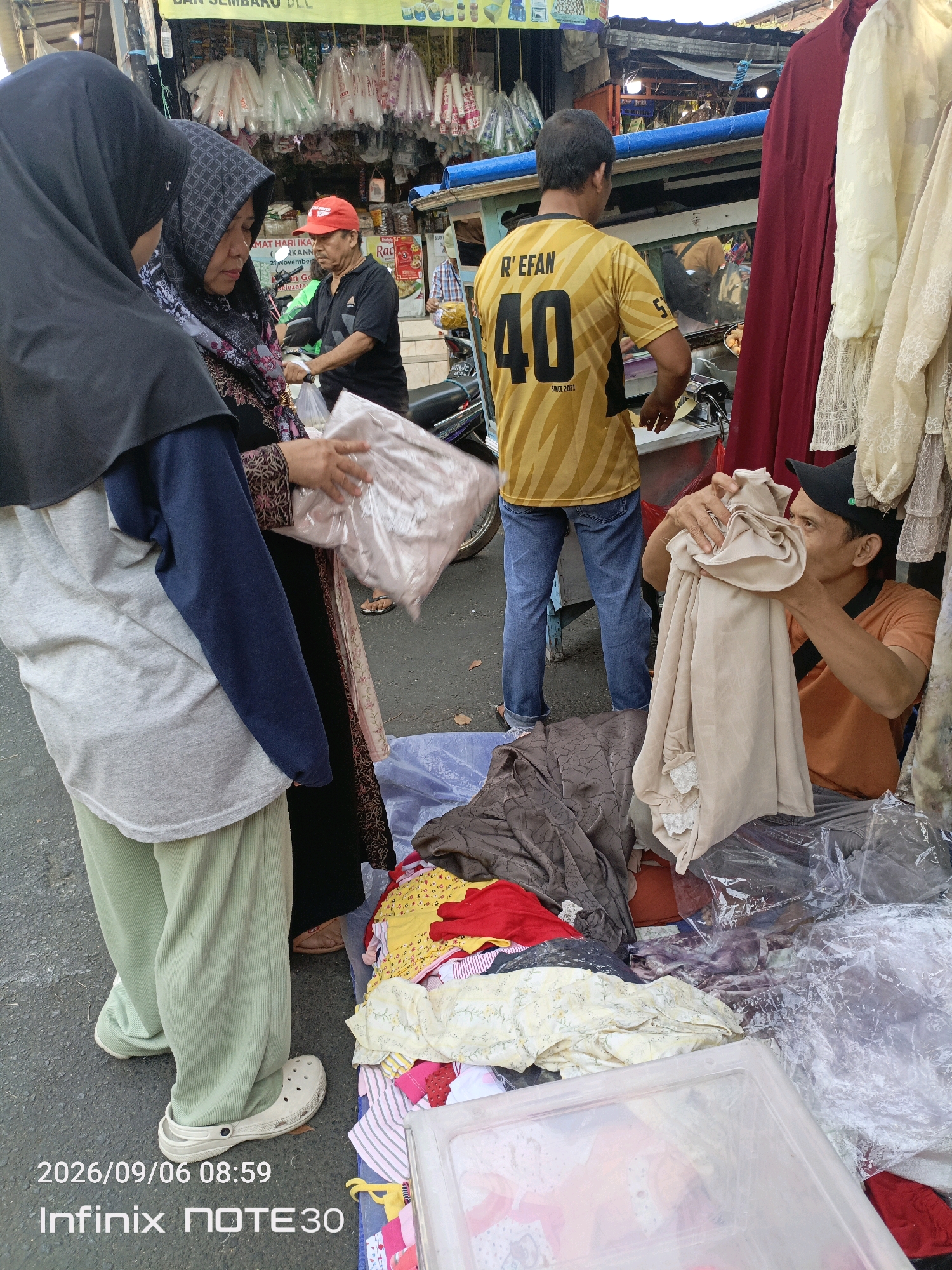 The image depicts a street scene in Central Jakarta, Indonesia, where a street vendor is arranging clothing. In the foreground, three individuals are visible. Two women, one wearing a black hijab and the other a patterned headscarf and dark clothing, are looking at a stack of clothing. The woman with the patterned headscarf is holding a transparent plastic bag filled with light-colored fabric. To their right, a man wearing an orange t-shirt and a black cap is examining a piece of beige fabric, appearing to be a vendor. Behind them, another man in a yellow t-shirt with the number "40" printed on it and blue jeans stands near a cart. The environment suggests a bustling street market, with stalls and various items for sale, including clothing and what appear to be plastic bags hanging in the background. The ground is paved, and there are stacks of folded clothing and plastic containers on the ground. The timestamp in the lower left corner reads "2026/09/06 08:59" and "Infinix NOTE30".