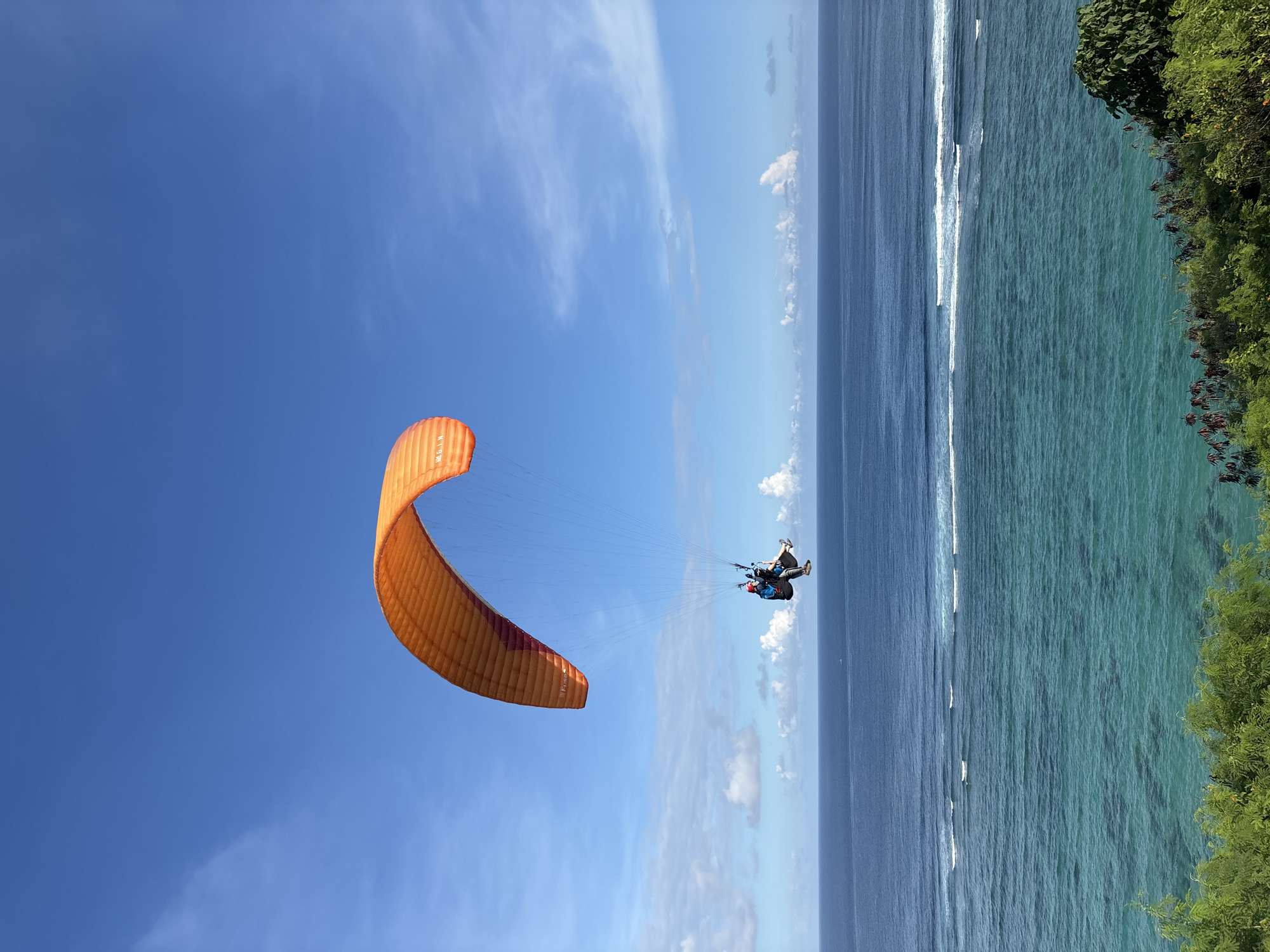 A paraglider with two occupants is soaring over the ocean near a coastal cliff in Kuta Selatan, Indonesia. The paraglider is predominantly orange with a red accent. The sky is blue with scattered white clouds. The ocean transitions from a darker blue further out to a turquoise hue closer to the shore, where white-capped waves are visible. Lush green vegetation covers the edge of the cliff.