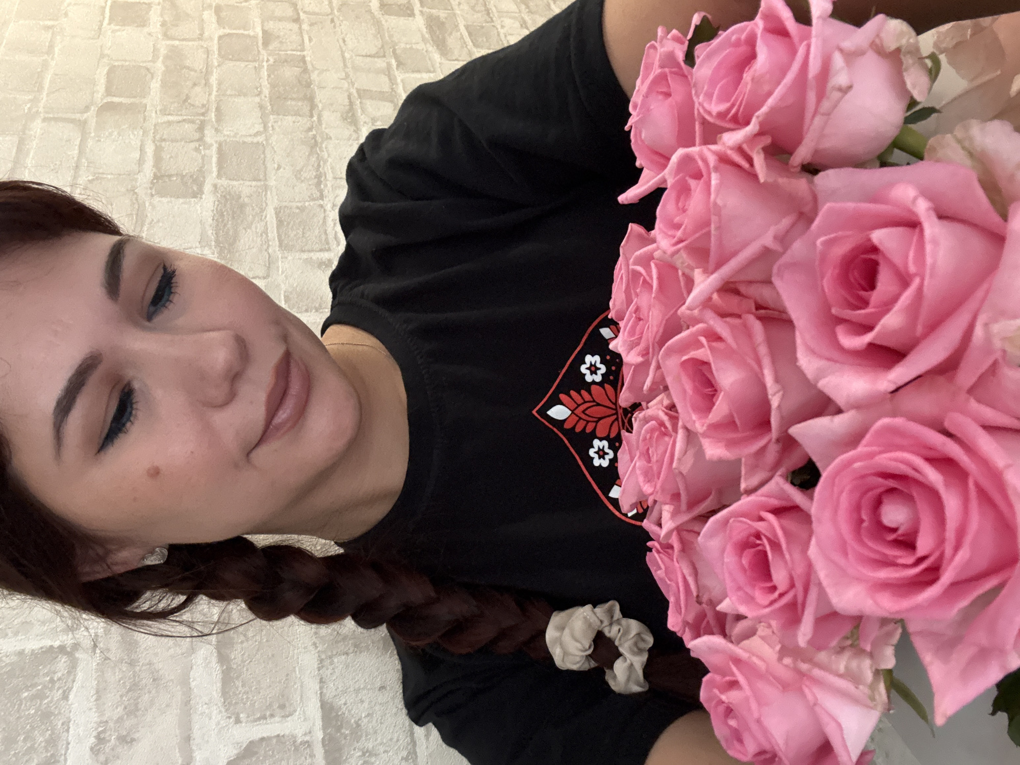 A young woman with dark hair braided down her back and a black t-shirt is holding a bouquet of pink roses. She is looking down at the flowers with a gentle smile. The background is a textured, light-colored brick wall. The lighting is soft and natural.