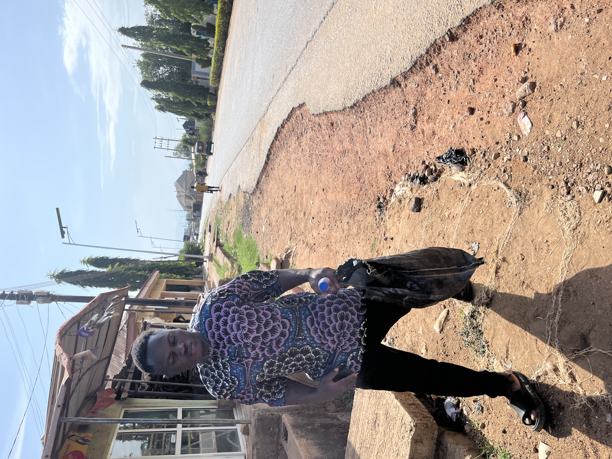 A man stands near a road in Jos, Nigeria. He is wearing a patterned shirt and black pants, holding a blue water bottle in one hand and what appears to be a large, dark sack in the other. He is standing near a concrete structure, possibly a curb or low wall, with a building visible behind him. The road is cracked and the shoulder is littered with trash. Other buildings and trees can be seen in the distance under a clear sky.