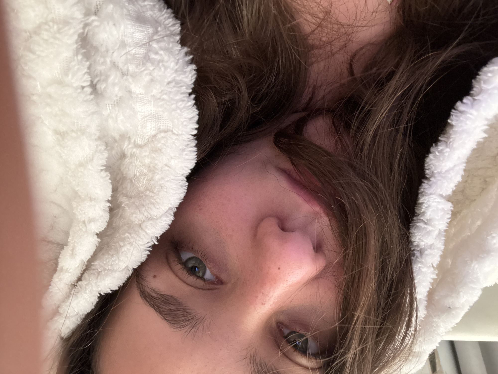 A close-up, high-angle selfie captures the face of a young individual, likely female, with fair skin, dark brown wavy hair, and green-hazel eyes. The person's head is tilted, presenting an inverted perspective, and they gaze directly into the camera with a neutral, relaxed expression.
They are partially enveloped in a soft, fluffy white garment, possibly a robe or blanket, which frames the left side of their face and head, contributing to a sense of comfort. Strands of their brown hair fall casually across their face and forehead. Visible skin details include a small mole near the bridge of the nose and another on the right cheek.
The lighting is soft and diffused, characteristic of an indoor setting, without harsh shadows. The background is out of focus and indistinct, suggesting a private, relaxed environment such as a bedroom or living space. The image conveys a moment of candid relaxation. This scene is taking place in Milan, Italy. There is no visible text.