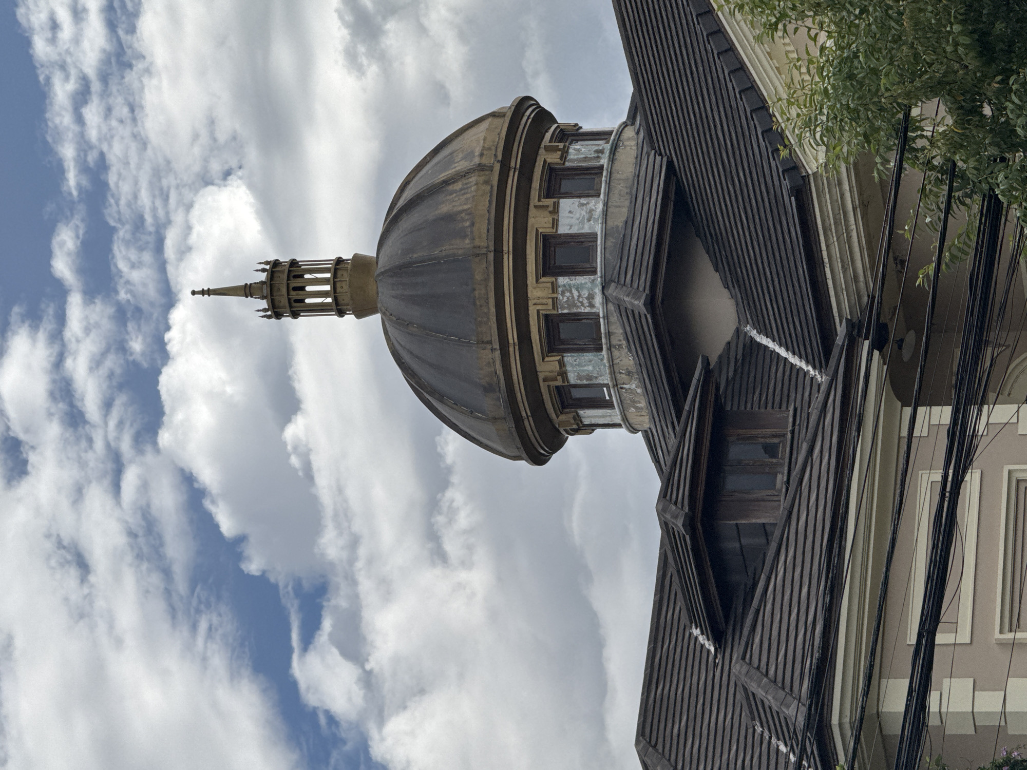 The image shows a close-up view of the dome and roof of a building in Da Lat, Vietnam, under a cloudy sky. The dome, made of dark metal with a golden patina, is topped with a decorative spire. Below the dome, a circular section of the building features several square windows. The roof is tiled in dark grey, with angled sections and dormer windows. The lower part of the building visible is painted a pale pink with white trim. Electrical wires run horizontally across the foreground, obscuring some of the lower facade. Green foliage from a tree is visible on the right side of the frame. The overall impression is of an older, possibly historic, building with architectural elements that suggest a European influence, common in Da Lat.