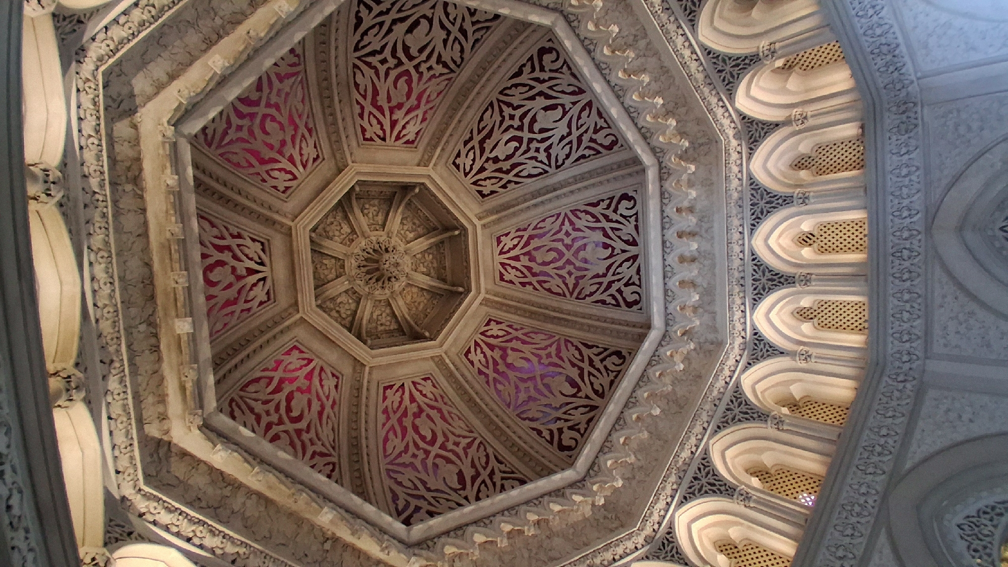 The image shows the interior of the Pena Palace in Sintra, Portugal. The viewpoint is looking directly upwards at an octagonal dome. The dome is elaborately decorated with carved stonework and a vibrant red textile with intricate patterns visible in the recessed sections. The lower part of the structure features a series of arched windows with decorative latticework, interspersed with ornate architectural details. The lighting appears to be a mix of natural light coming through the windows and artificial interior lighting, casting subtle shadows and highlighting the textures of the architecture.