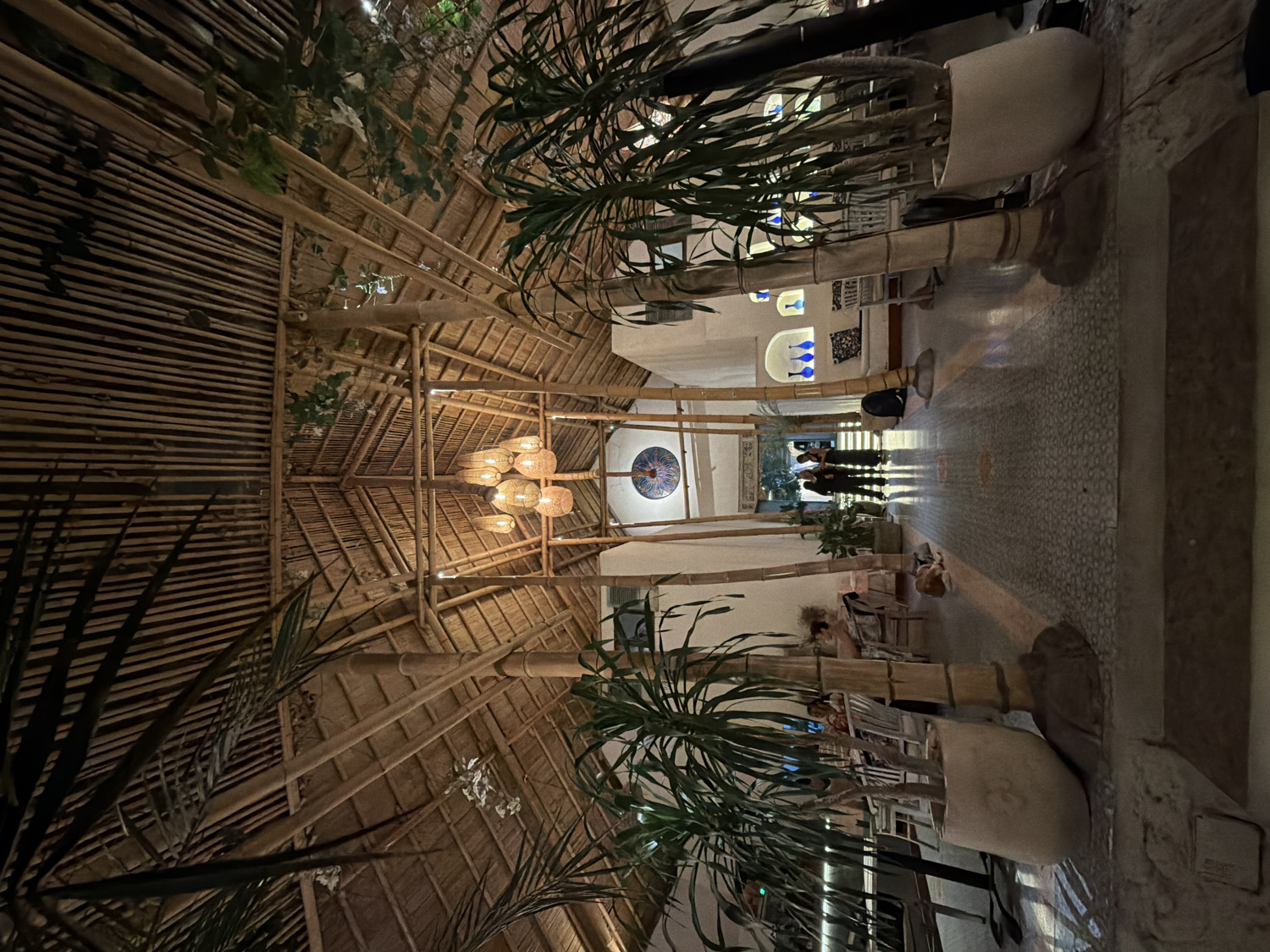 The image captures the interior of a spacious, tropical-themed establishment, likely a restaurant or lounge, at night or in the evening. The dominant feature is the high, vaulted ceiling constructed from an intricate lattice of bamboo poles and what appears to be a thatched or woven material, creating a rustic and natural aesthetic. Several decorative chandeliers, fashioned from woven materials, hang from the ceiling, casting a warm, inviting glow that illuminates the space.
The scene depicts a long, grand corridor or central aisle leading into the distance. Along both sides of this pathway are large, leafy potted plants, adding to the lush, outdoor-inspired ambiance. The floor is covered with a decorative patterned tile, guiding the eye towards the far end of the corridor.
At the distant end of the aisle, several people are visible, silhouetted against a lighter background, suggesting an entrance to another area or an outdoor space. Their exact actions are indistinct, but they appear to be standing or walking. On the left side of the corridor, near the foreground, furniture like tables and chairs are visible, with at least one person seated, indicating dining or lounging areas. The walls along the corridor feature decorative elements, including arched niches with blue vases and a circular, colorful mosaic-like art piece.
The warm, artificial lighting from the chandeliers and other unseen sources indicates that the photograph was taken after dusk. The architectural style, use of natural materials like bamboo and thatch, and abundant greenery strongly suggest a resort or upscale casual dining setting, consistent with the specified location of North Kuta, Indonesia. No discernible text is visible in the image.