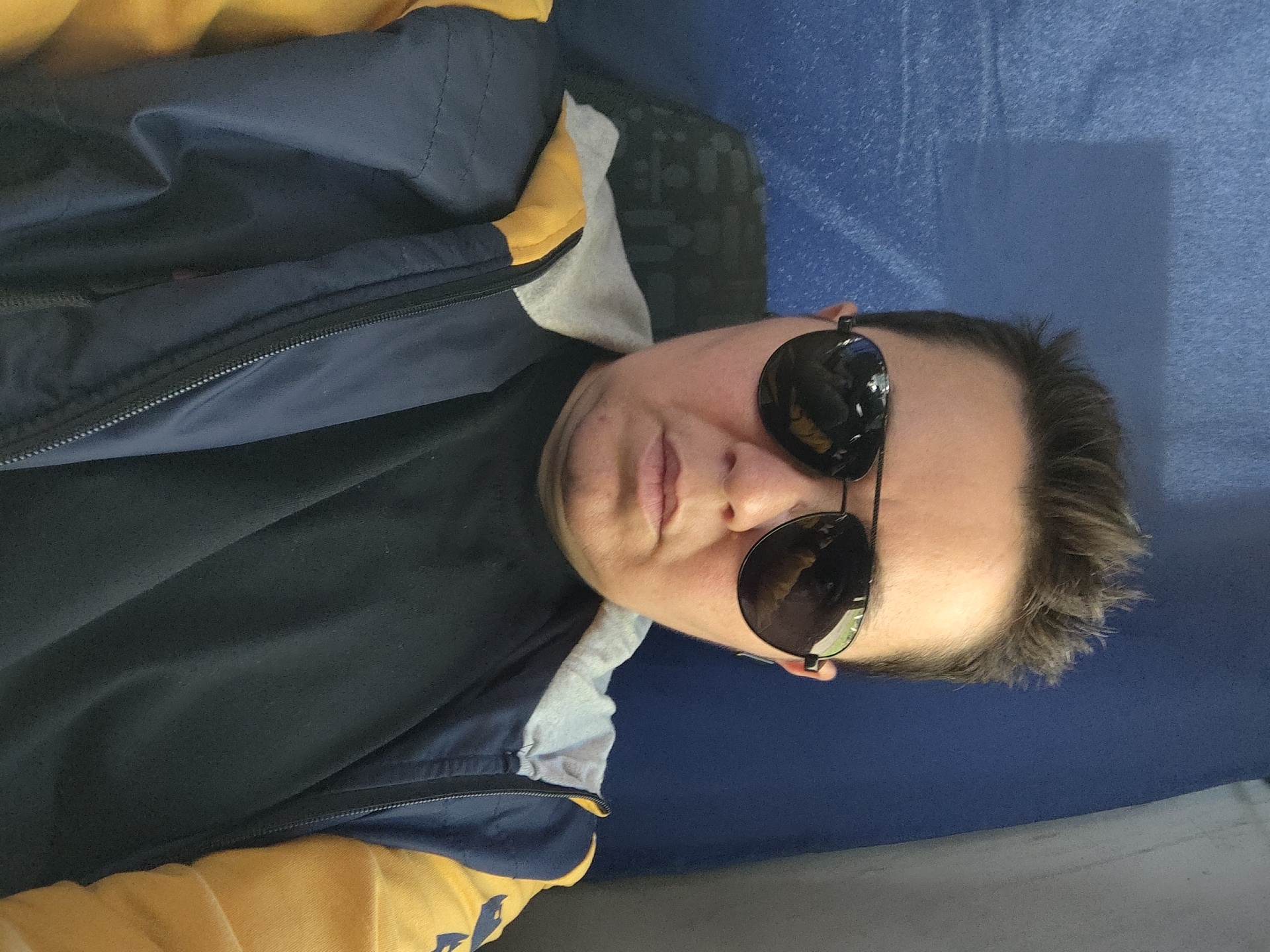 The image shows a close-up selfie of a person wearing aviator sunglasses, a black t-shirt, and a navy blue and yellow jacket. The person has short, dark, spiky hair. The background is a textured blue surface, possibly a wall or upholstery. The lighting suggests an indoor environment. The scene does not provide any specific information to indicate a location in Kratovo, Russia.