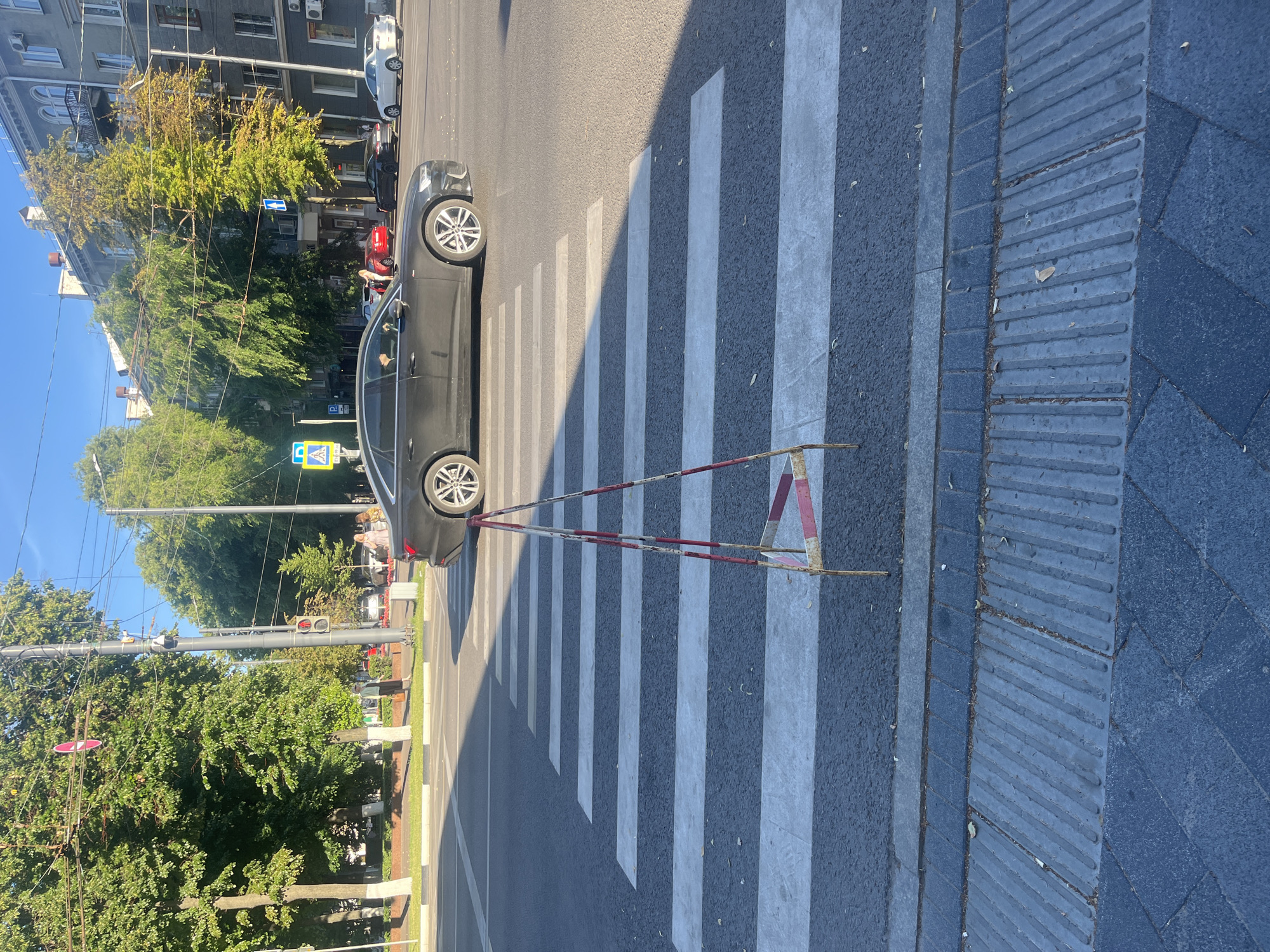 This media file captures a street scene in Novaya Usman, Russia, likely during a sunny day given the strong shadows. The primary subjects are a black car crossing a pedestrian street, also known as a crosswalk, and a metal barrier with red and white markings positioned at the edge of the sidewalk. The crosswalk features prominent white stripes painted on asphalt. The sidewalk itself is composed of different textured paving stones, with a section exhibiting raised ridges, possibly for tactile paving.
In the background, trees with green and yellowing leaves suggest it might be late summer or early autumn. Buildings line the street, some with windows and balconies visible. Traffic lights are also present, indicating an active intersection. A blue directional sign with a white arrow and a yellow diamond-shaped sign with a pedestrian symbol are visible, further reinforcing the context of a pedestrian crossing.
The activity is focused on the car navigating the crosswalk. The scene appears to be a typical urban street setting, devoid of any strong emotional cues. The lighting and shadows suggest the sun is at an angle, likely mid-morning or late afternoon. There is no visible text in the image, other than on the traffic signs which are too small to read.