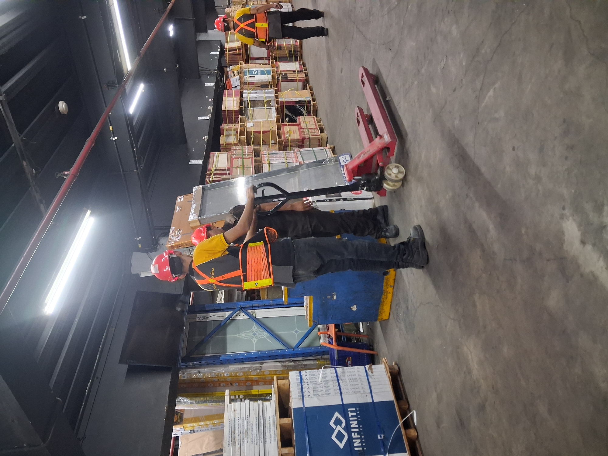 The image depicts the interior of a warehouse in Depok, Indonesia, filled with stacked merchandise, primarily tiles in blue and white packaging labeled "INFINITI GRANITE TILE." A red manual pallet jack is visible in the foreground, with two individuals wearing red hard hats and high-visibility vests actively engaged in moving a large, sheet-like object. One individual is operating the pallet jack, while the other stands nearby, seemingly assisting. The warehouse environment is characterized by concrete flooring, exposed ceiling structures with industrial lighting, and rows of wooden pallets laden with goods. The overall scene suggests a typical operational day in a logistics or distribution facility.