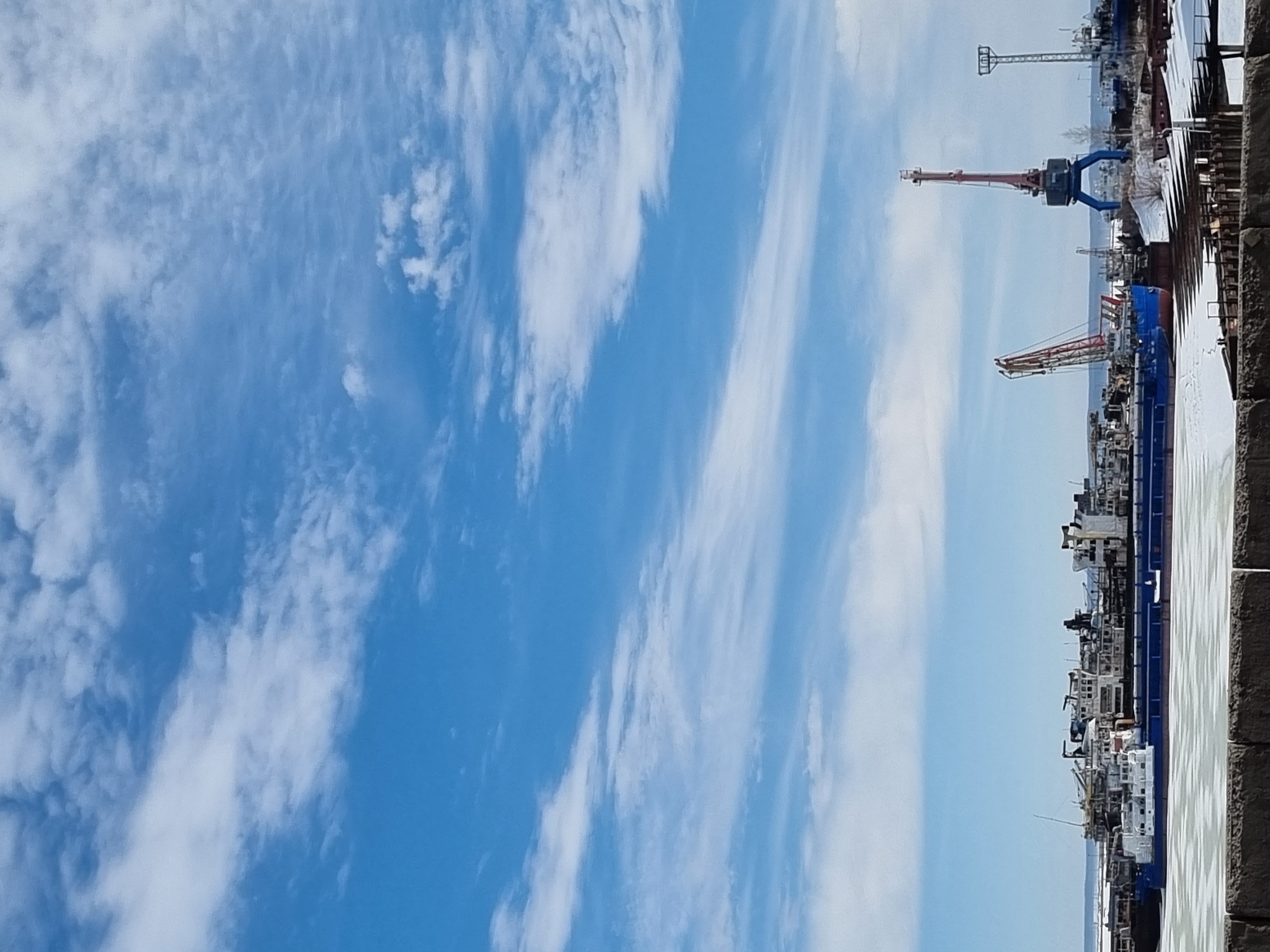The image presents an industrial waterfront scene in Chaikovsky, Russia, observed during daylight under a bright blue sky with scattered white clouds. In the immediate foreground, a dark, textured barrier, possibly concrete or stone, is visible, partially topped with snow or ice. Beyond this barrier, a body of water, likely a section of the Kama River or Votkinsk Reservoir, is largely covered in extensive sheets of white and light blue ice, indicating winter conditions.
Multiple large vessels are moored within this ice-laden water. These vessels are predominantly industrial barges or cargo ships, characterized by dark blue hulls and white superstructures, with various cranes, masts, and deck equipment visible. One prominent blue hull is situated to the left of the image center, with a smaller white vessel positioned in front of a larger blue one.
On the distant shoreline, further industrial infrastructure is present. A tall, red and blue gantry crane with a high boom stands on the right side of the frame. To its left, another crane, featuring a red and white boom, is visible. Further right, a tall metal utility pole or light tower is also present. The ground on the shoreline exhibits snow cover and some sparse, bare trees or shrubs. No individuals or specific actions are discernible within the scene.