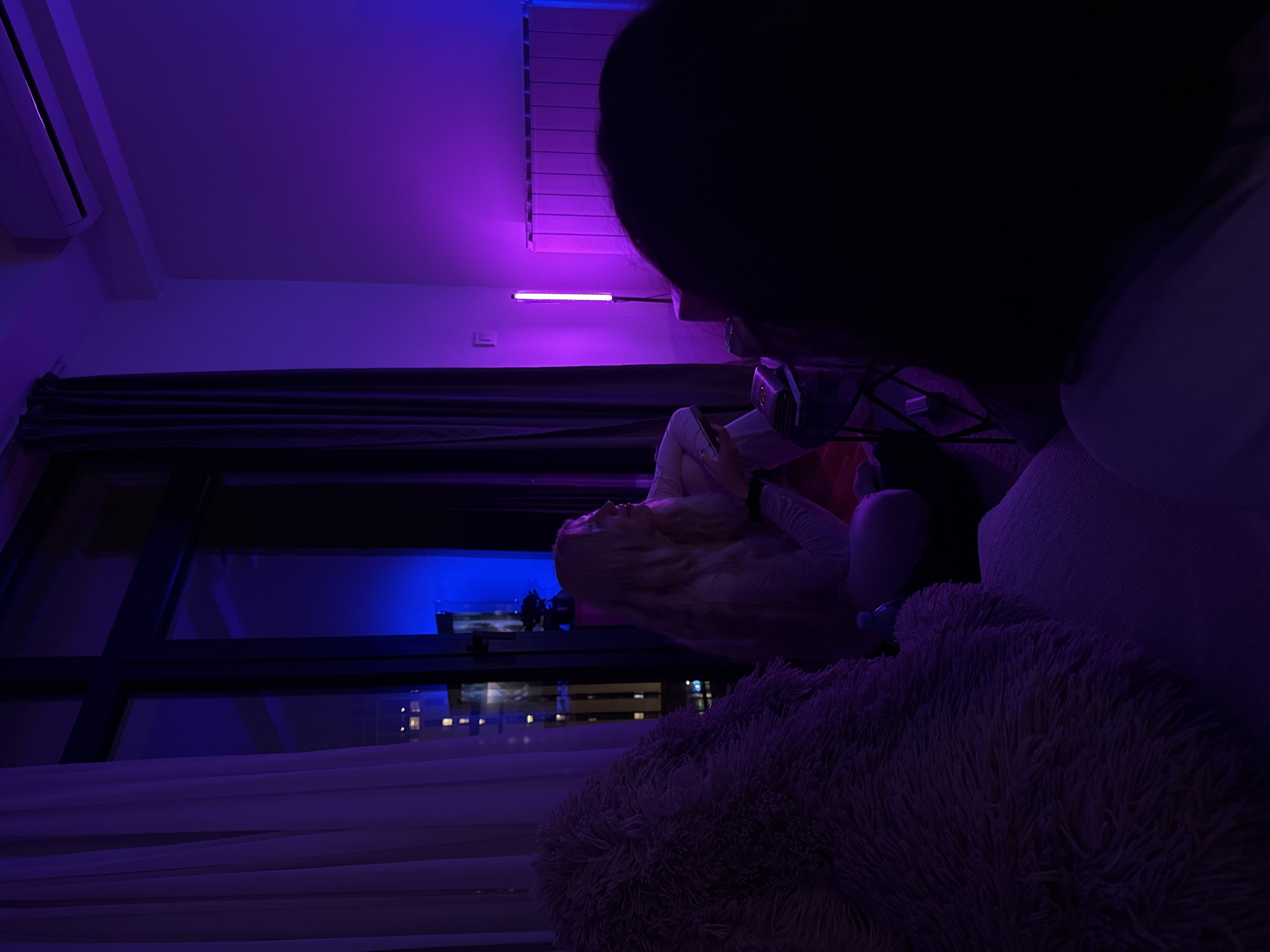 A person with long blond hair is lying down indoors near a large window with open curtains. The room is illuminated with purple and blue lighting. Outside the window, city lights are visible, suggesting an urban environment. The presence of an air conditioning unit on the wall indicates a modern apartment setting. Another person with dark hair is positioned in the foreground, partially obscuring the scene. The room contains soft furnishings, including a fluffy cushion. The location is the city of Belgrade, Serbia.