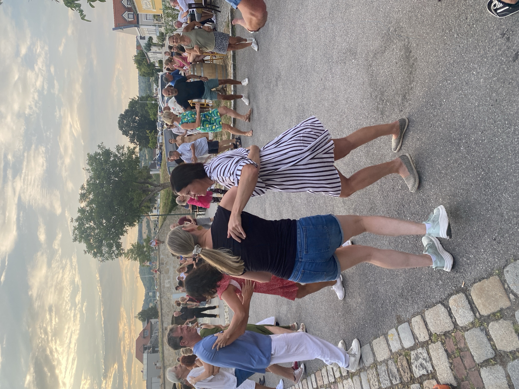 This is a medium shot, eye-level, outdoor photograph taken in Purbach, Austria, depicting a group of people dancing in what appears to be a street or plaza area. The sky is filled with dramatic clouds during what seems to be sunset.
In the foreground, two couples are dancing. On the left, a man in a blue t-shirt and white pants is dancing with a woman in a red, polka-dot dress. To their right, a woman in a black t-shirt and denim shorts is dancing with a man in a black shirt and shorts. Another woman in a black and white striped dress is dancing with a man whose arm is visible on the far right edge of the frame.
Behind the dancing couples, a larger group of people are socializing and observing. Some are seated at tables on the right side of the image. The background shows buildings with tiled roofs, trees, and a hilly landscape. The ground is a mixture of cobblestones and asphalt. The overall atmosphere is festive and casual.