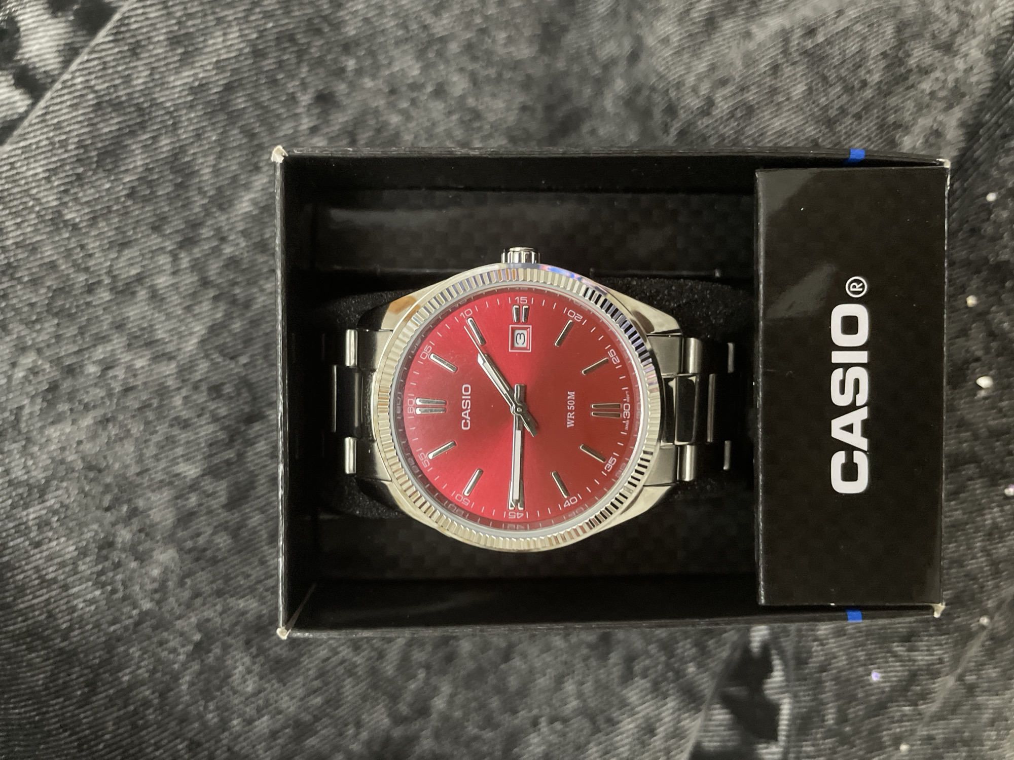 A silver-toned Casio wristwatch with a vibrant red dial is prominently displayed within its black presentation box. The watch features silver hour markers, luminous hands, and a date window at the 3 o'clock position showing the number '3'. Around the outer edge of the dial, a minute track is visible with Arabic numerals marking every five minutes, from '05' to '55'. The watch dial also bears the text "CASIO" and "WR 50M", indicating its water resistance. The watch case has a distinct fluted bezel, and it is fitted with a matching silver-toned stainless steel bracelet. The watch is cradled in a black foam insert within the rectangular black box. The side of the box is visible, featuring the brand name "CASIO®" in white lettering. The background consists of a dark, textured fabric, likely denim or a similar textile, which provides a neutral backdrop for the watch and its packaging. The image captures a static display, possibly an unboxing or product shot. This scene takes place in Ihtiman, Bulgaria.