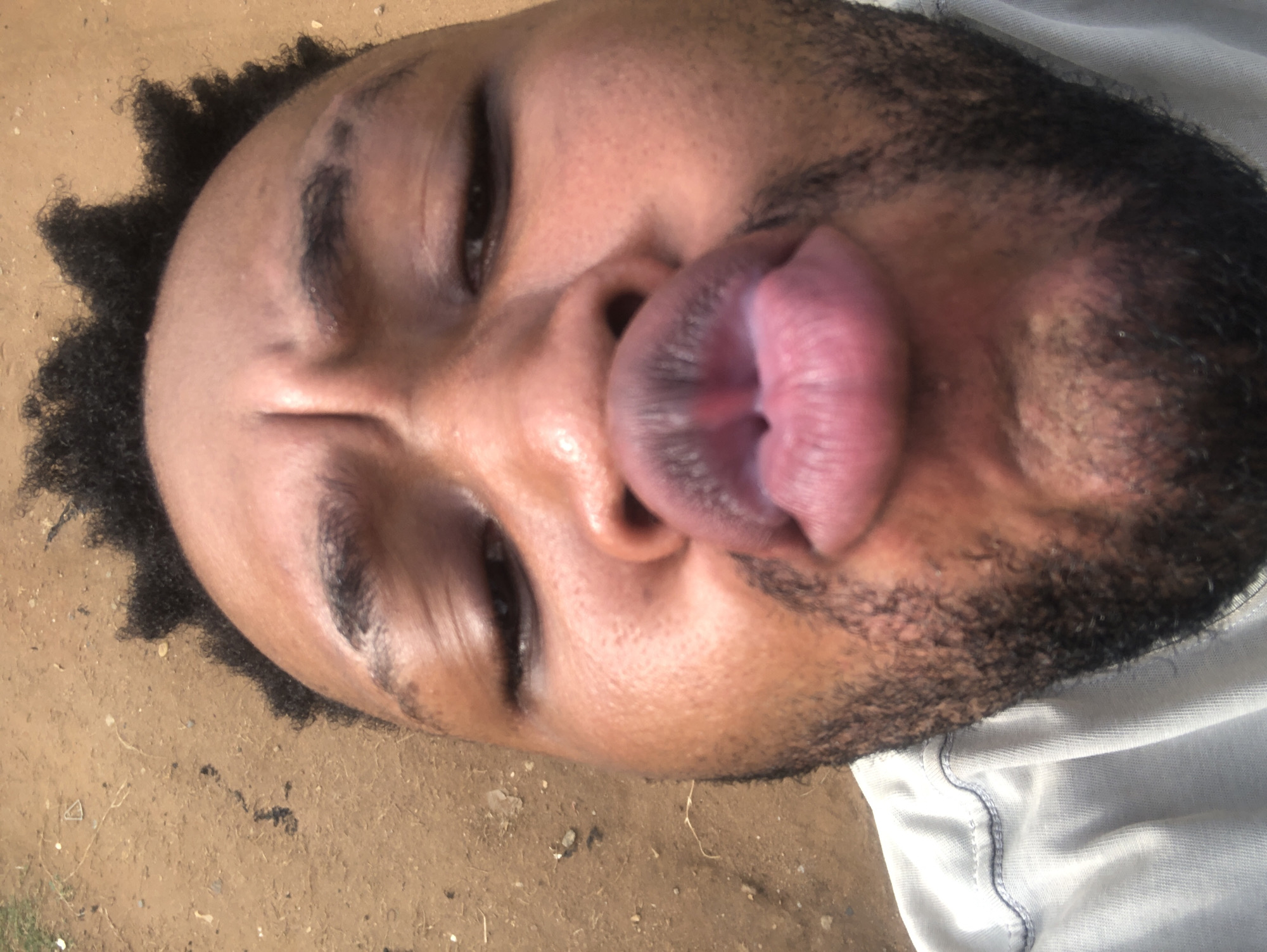 A close-up portrait of a man's face, taken at an extreme angle. The man has dark hair and a beard. His lips are pursed outwards. He is lying on a surface that appears to be dirt. Part of a light-colored garment is visible in the lower right corner. The location is unspecified.