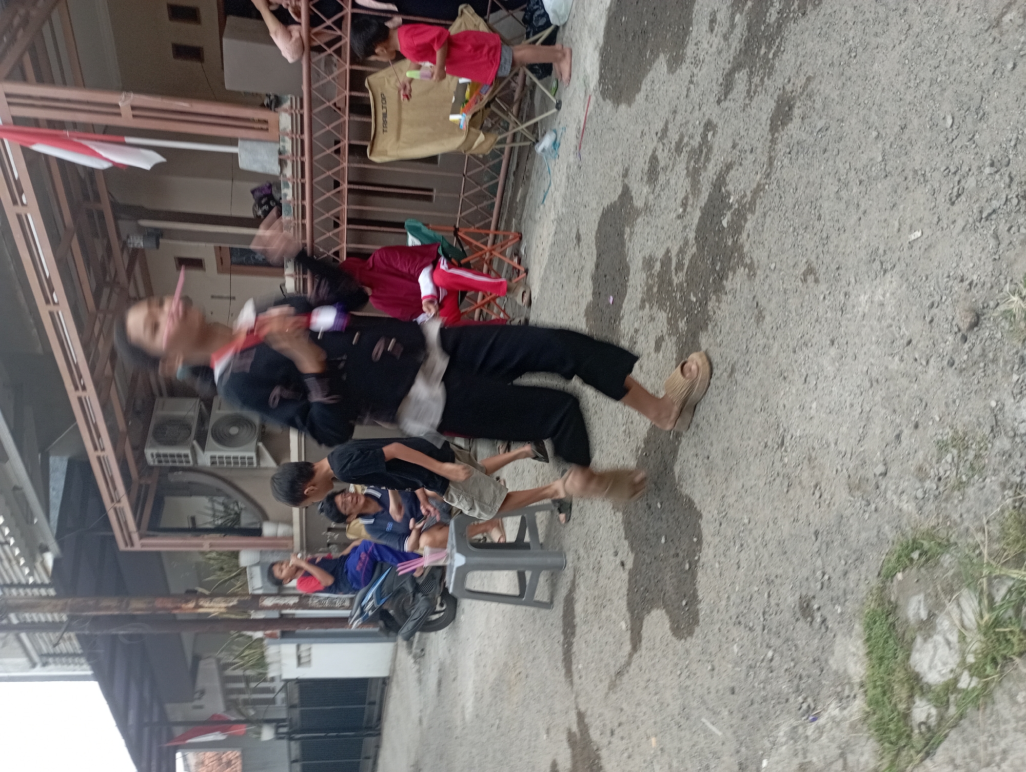 In a residential area of Purwakarta, Indonesia, a group of people are gathered outdoors. The focus is on a young person in the foreground, blurred by motion, with what appears to be a pink object held to their mouth, possibly engaged in a game or activity. To their left, a grey plastic stool holds a pink cup with several pink straws. Behind this, a motorcycle is parked, and several children are visible sitting or standing. Further in the background, other individuals are seated on folding chairs. The environment is a paved street with some patches of grass. Buildings and a fence are visible in the background. A red and white Indonesian flag is also displayed.