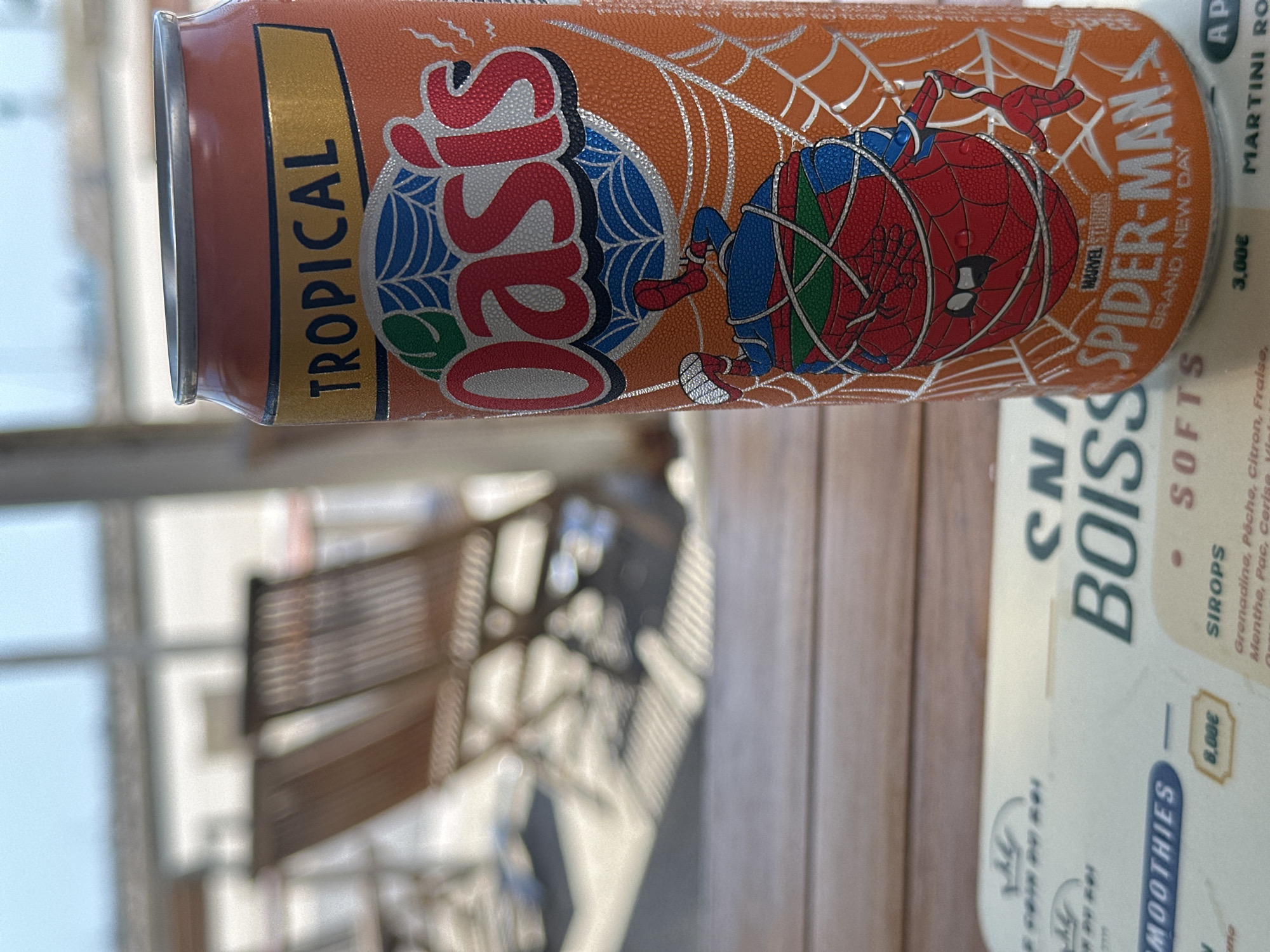 An outdoor, eye-level, close-up shot shows an orange aluminum can of Oasis Tropical Spider-Man Brand New Day drink. The can is covered in condensation and is positioned on a wooden table. Behind the can, two blurred wooden chairs and a white building are visible. The can's label features the Oasis logo with a web design and an illustration of Spider-Man entangled in webbing. A blurred menu is visible in the foreground, showing text including "Smoothies," "Boissons," "Softs," and "Sirop." The lighting suggests it is daytime.