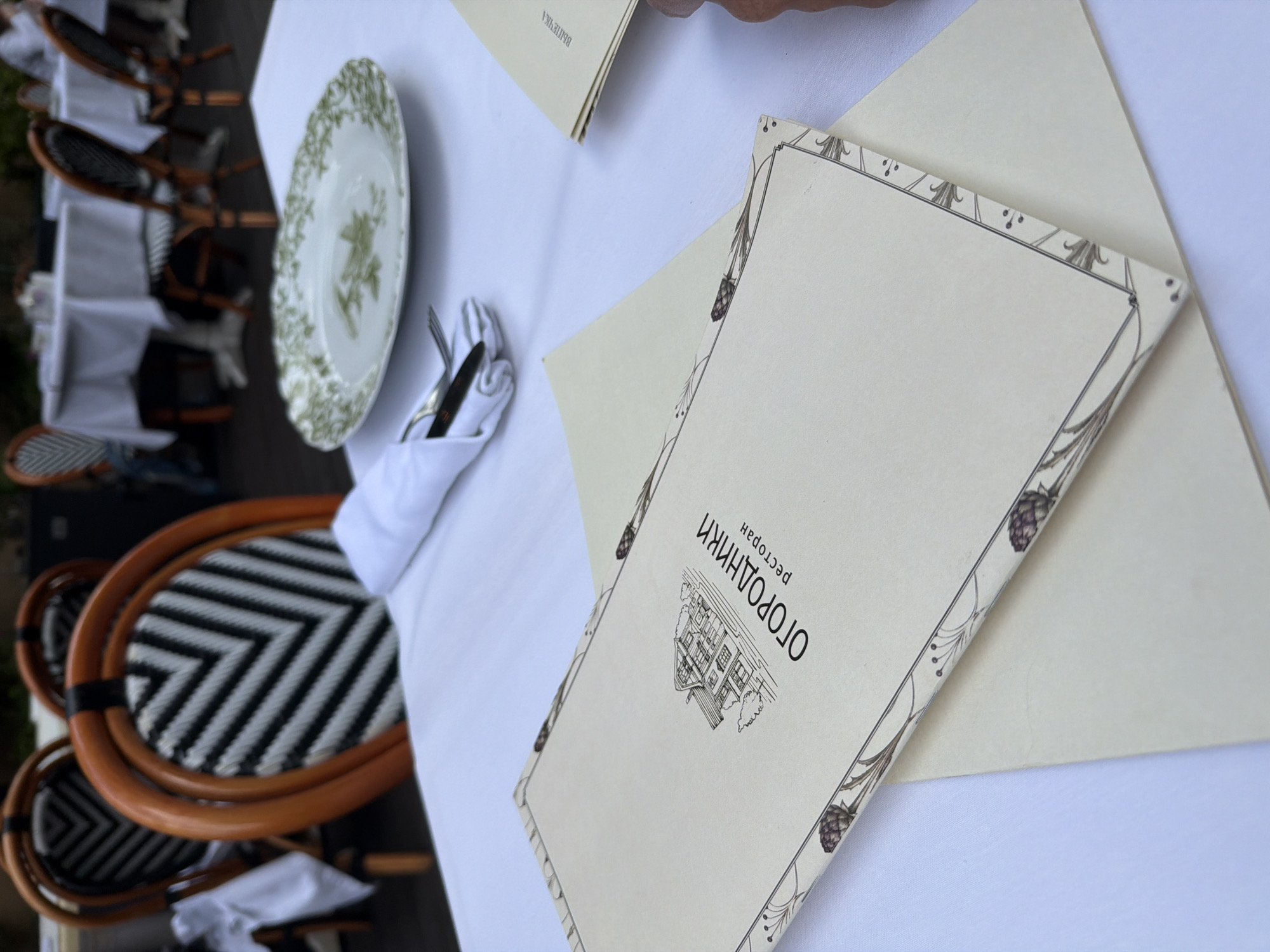 An outdoor restaurant setting in Saint Petersburg, Russia is depicted, featuring a white-clothed table with place settings. A menu with the text "ОГОРОДНИКИ ресторан" is prominent, featuring a drawing of a house and decorative botanical borders. A plate with a green floral design, silverware wrapped in a white napkin, and another menu visible in the background are also present. The chairs have a distinctive woven design with black and white patterns and wooden frames. The overall ambiance suggests a pleasant dining experience.