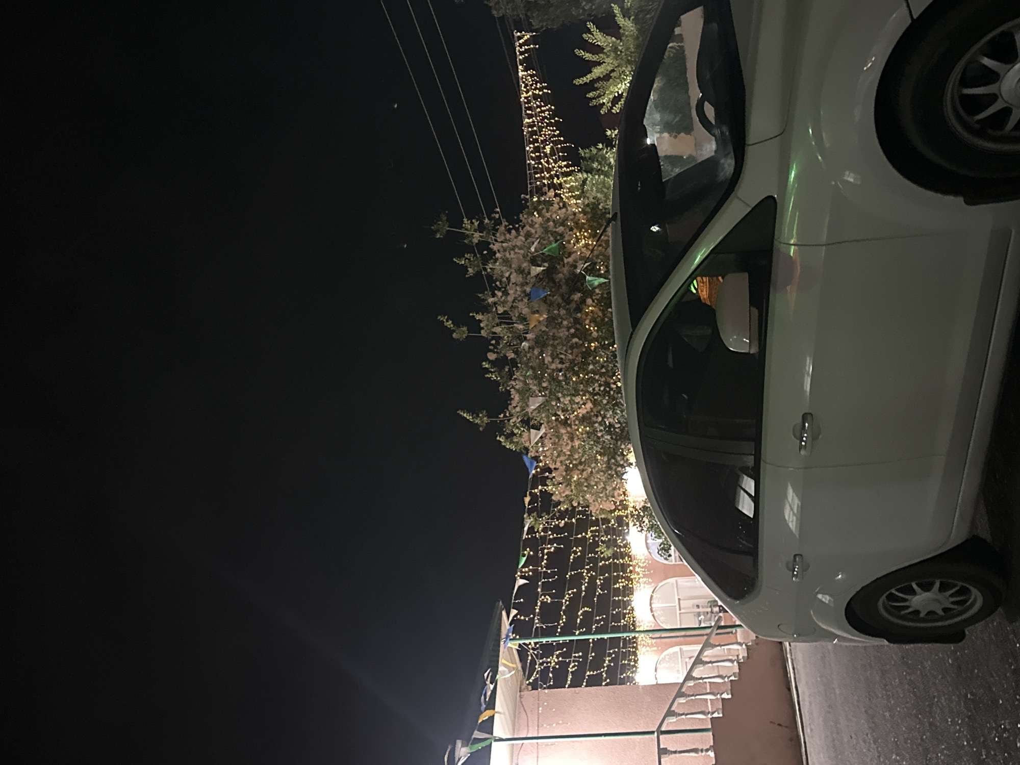 The image shows a white car parked at night, possibly in Subutli-Miatli, Russia. The car is viewed from the side, with its rear wheel visible. The scene is illuminated by strings of small, warm-toned lights that are draped across trees and a building in the background. There are also small triangular flags, some of which appear to be green, blue, and yellow, strung along with the lights. The sky is dark, indicating it is nighttime. The building in the background has arched windows, and a railing is visible in the foreground. The overall atmosphere appears festive and celebratory, possibly due to the decorations. There is no visible text in the image.