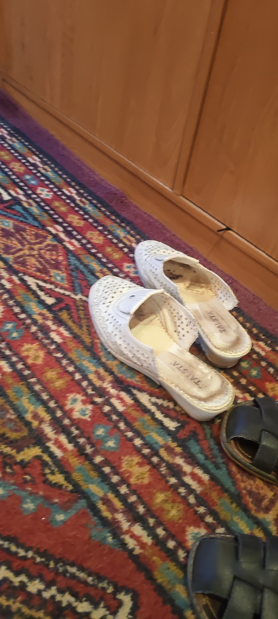 The image shows a pair of worn, white slip-on shoes with a perforated design and a pair of dark-colored sandals on a colorful, patterned rug. The shoes appear to have some dark staining on the insoles and the words "TALSTA" are visible on the inner sole of the right shoe. The sandals are simple, with thick straps. The rug has a complex geometric pattern with colors including red, blue, yellow, and black. In the background, a wooden surface, possibly a cabinet or door, is visible. The scene appears to be indoors, likely in a home setting. There are no people visible in the image. The lighting suggests it could be daytime, but no specific time can be determined. The location context provided is Mezhdurechensk, Russia. The overall impression is one of everyday domestic life, with casual footwear placed on a decorative rug.