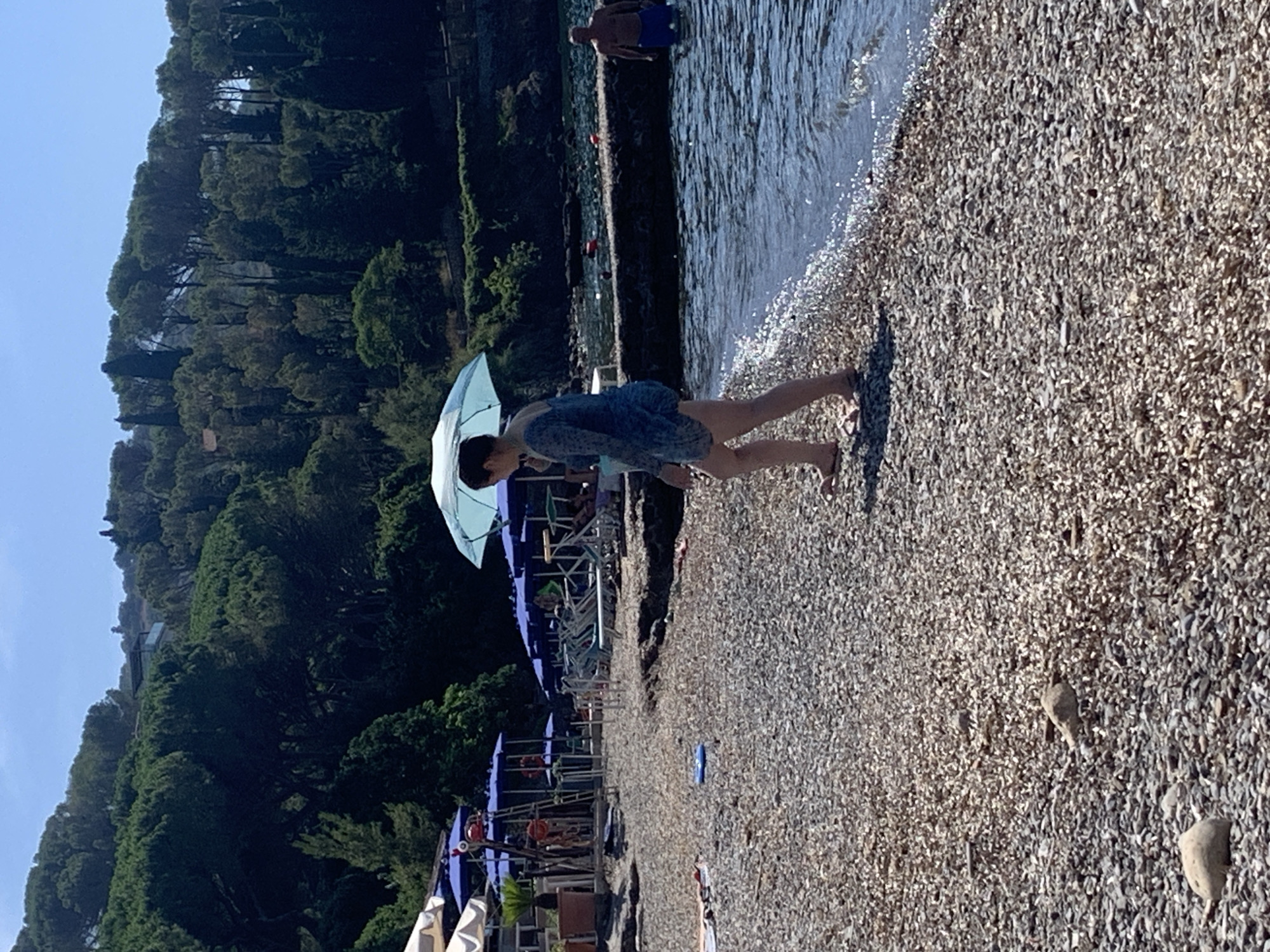 The image depicts a sunny day at a pebble beach in Pozzarello, Italy. A woman, wearing a light blue, sheer, knee-length dress and flip-flops, walks along the water's edge, holding a light blue umbrella over her head. Her dark hair is pulled back. In the background, a man stands in the water wearing blue swim trunks. The water is clear and gently laps at the shore. The beach is covered in small, light-colored pebbles. To the left of the image, there are rows of blue umbrellas and lounge chairs, suggesting a populated beach area. A lush green hillside covered in trees rises behind the beach. The sky is clear and blue.