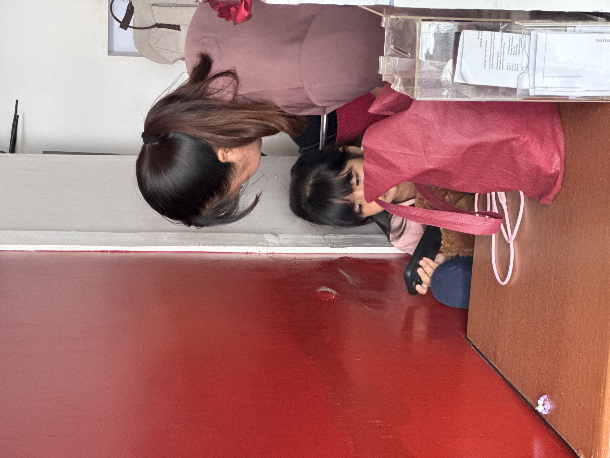A woman with long brown hair is interacting with a young girl who is peering out of a red tote bag. The woman is wearing a light brown sweater and the girl is wearing a patterned brown and pink shirt. They are situated next to a brown wooden surface with a clear plastic container on top, which contains papers. The background consists of a wall, and the floor is red. The location is Kuta Selatan, Indonesia.