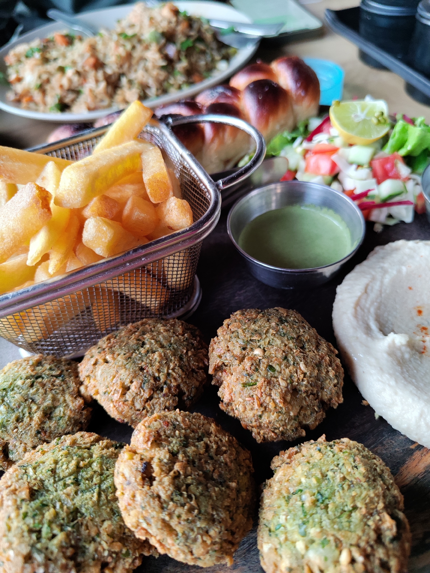 This is a close-up, high-angle shot showcasing a diverse spread of food items, likely a meal at a restaurant or cafe. The scene is well-lit, highlighting the textures and colors of the dishes.
In the immediate foreground, several pieces of falafel are visible, characterized by their textured, golden-brown and green surfaces, indicating a mix of ingredients. To the right of the falafel is a generous portion of creamy white hummus, lightly garnished with a red spice.
Central to the composition, behind the falafel, is a metal mesh basket overflowing with golden-yellow french fries. Adjacent to the fries, two small, shiny metal bowls hold dips; one contains a vibrant green sauce or dip, while the other is partially visible.
Further back, a fresh side salad is arranged on the dark surface, featuring chopped cucumbers, tomatoes, lettuce, and thin slices of radish, topped with a bright yellow lemon wedge. Behind the salad, a braided, golden-brown bread, resembling challah or a similar type, is placed.
In the softly blurred background, a white oval plate holds a serving of fried rice, mixed with vegetables and possibly other ingredients. The overall setting appears to be an indoor dining environment, with a dark wooden or stone surface serving as the table or tray. There is no visible text in the image.