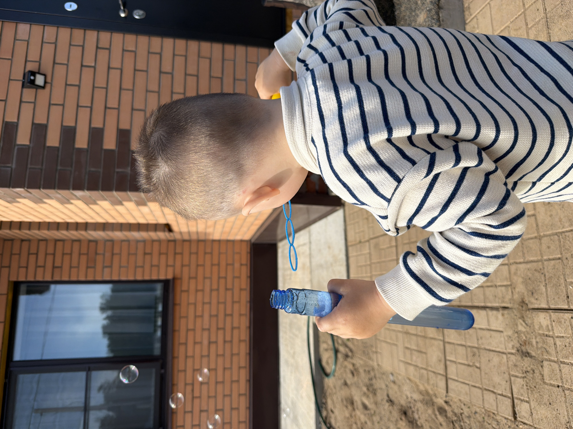 This image features a young child, viewed from behind, blowing soap bubbles outdoors in Chaikovsky, Russia. The child is wearing a white long-sleeved shirt with prominent navy blue horizontal stripes. They are holding a tall, translucent blue bottle of bubble solution in their right hand, with a blue bubble wand, connected by a string, positioned near their mouth. Several iridescent bubbles are visible floating in the air, indicating the ongoing activity.
The setting appears to be the side of a building, characterized by a textured brick wall featuring a mix of lighter and darker brown bricks laid in a staggered pattern. A large window with a dark frame is set into the wall, reflecting parts of the sky and surroundings. The ground beneath the child's feet is paved with rectangular tiles. The scene is brightly illuminated by natural sunlight, suggesting a clear and sunny day, likely in the morning or afternoon, given the crisp shadows. The child's head has short, closely cropped hair. No visible text can be discerned anywhere in the image. The overall impression is one of simple, joyful outdoor play.