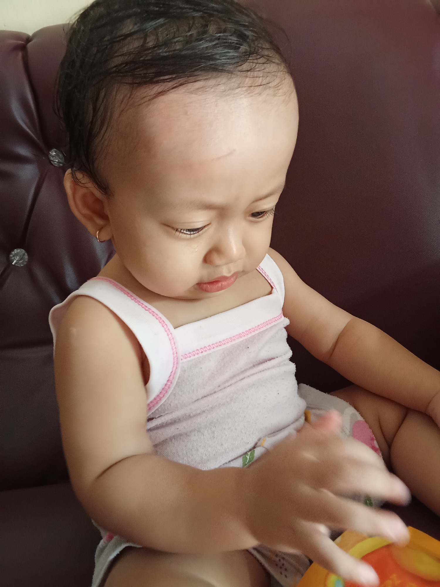 A close-up, high-angle shot shows a young toddler with dark hair and fair skin sitting on a brown leather sofa. The child is wearing a light pink tank top with white trim. The child's right hand is extended forward, and appears to be interacting with an object that is out of frame. The child's head is tilted down, and their gaze is directed towards their hand. The background is a blurred brown sofa. The image was taken in Tanah Mas, Indonesia.
